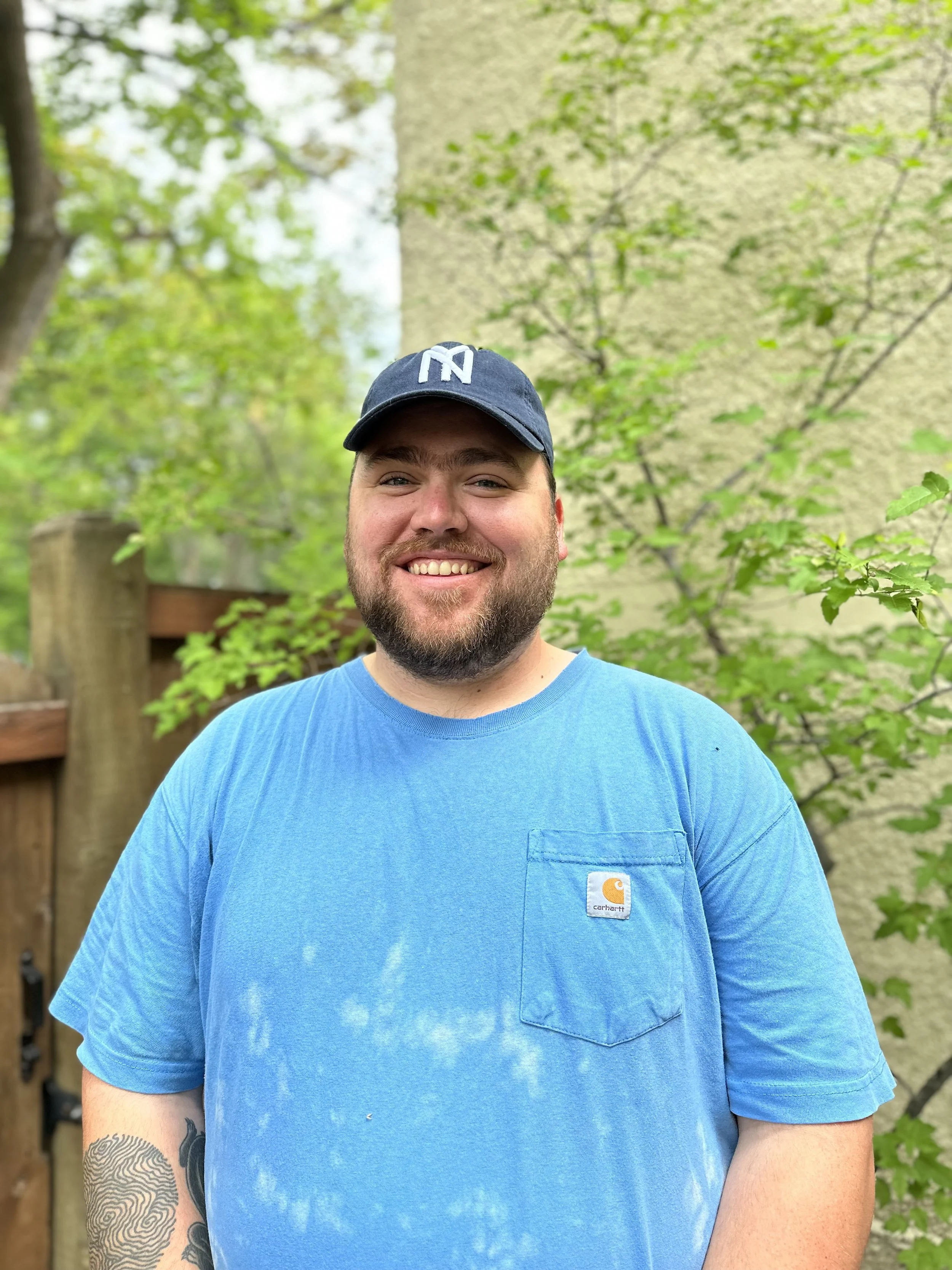 Image of Colter, a white man wearing a dark blue NY dad hat and a blue, short-sleeve carhartt shirt. Colter has a beard and and a sweet smile. 