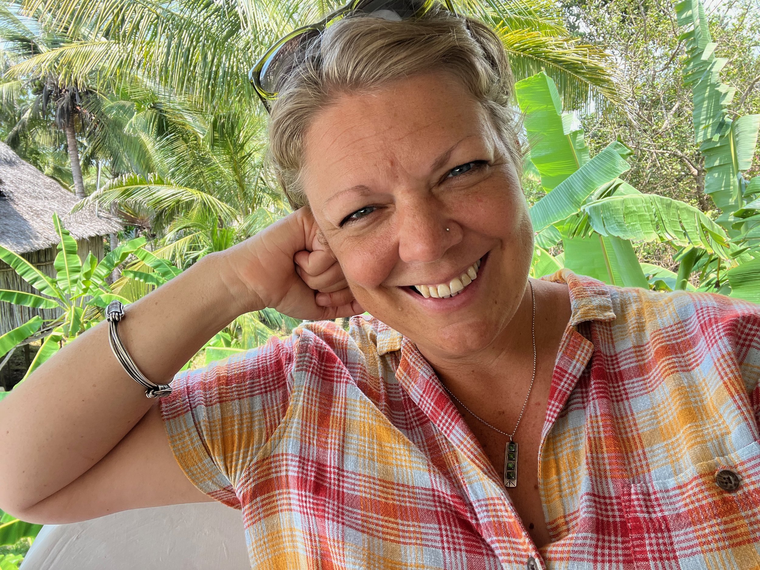 Photo of rachel, a white woman with blond hair, smiling at the camera while leaning on hand. She's wearing a plaid orange and pink blouse.