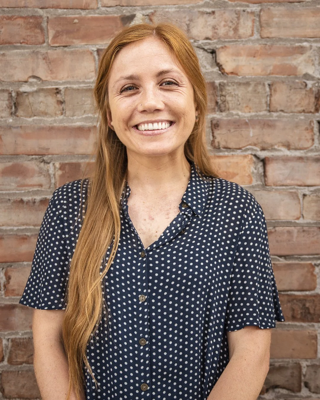 Image of Carlee, a white woman with red hair wearing a polka dot blouse. She smiles at the camera in front of a brick wall.