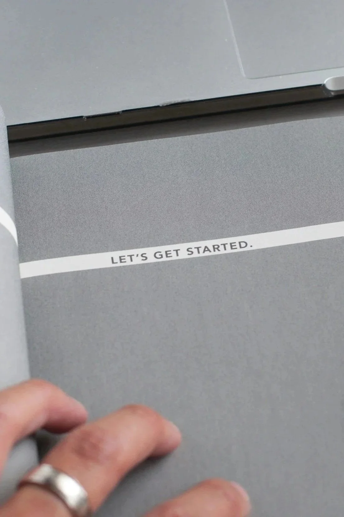 Close-up of a page in a booklet or notebook with a grey background, showing the words 'LET'S GET STARTED.' written on a white strip of paper.