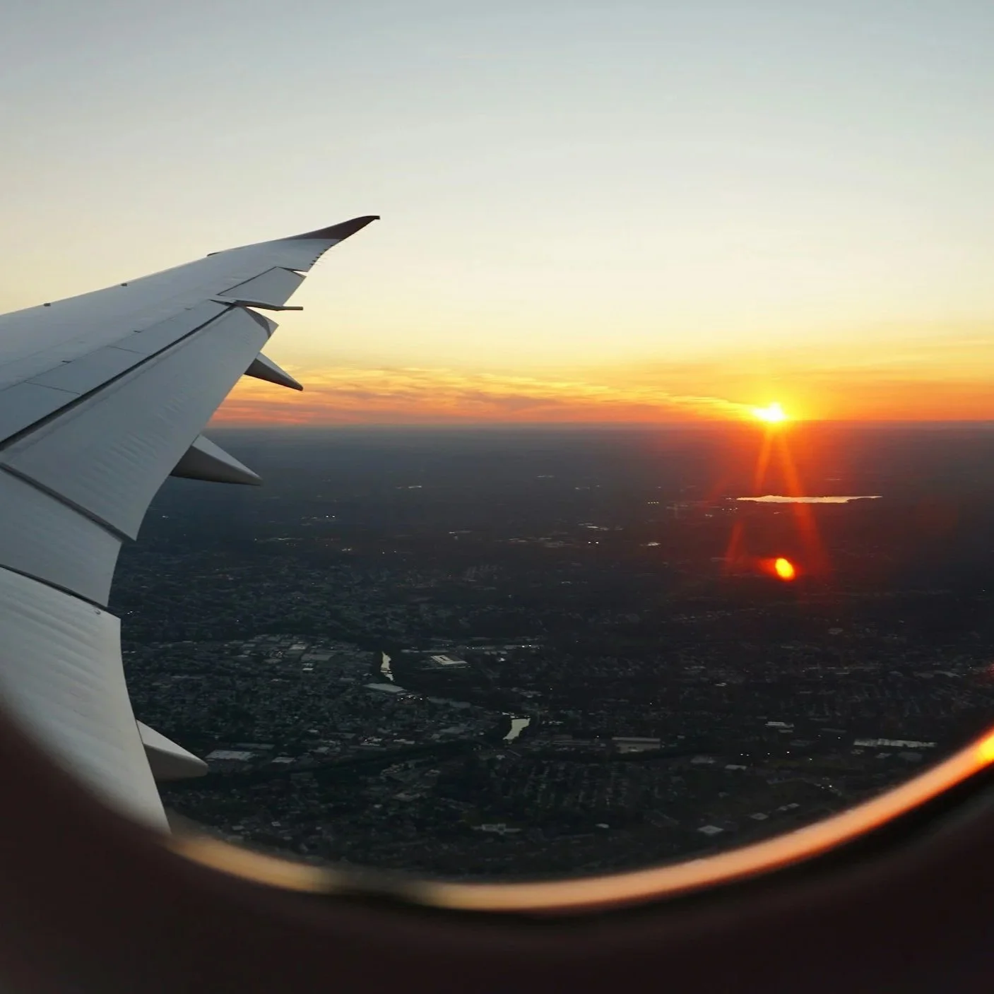 View of sunset from airplane window showing part of the wing and landscape below with a body of water.