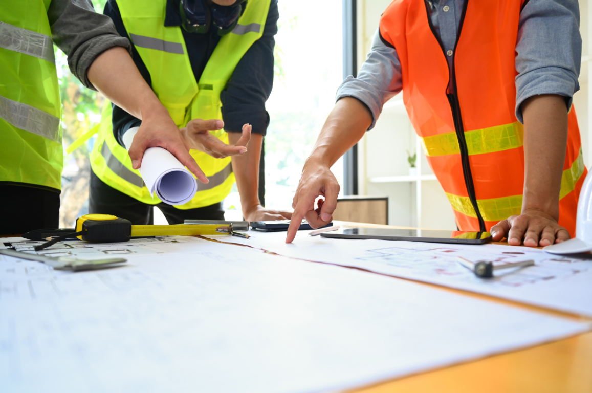 Two construction workers in safety vests discussing plans on a table, with tools and blueprints.