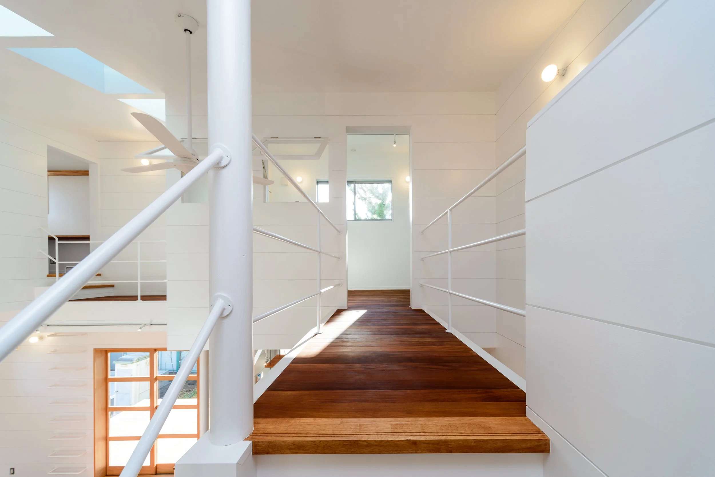 Interior view of a modern residential staircase with white walls, dark wooden steps, white metal railings, and large windows allowing natural light.