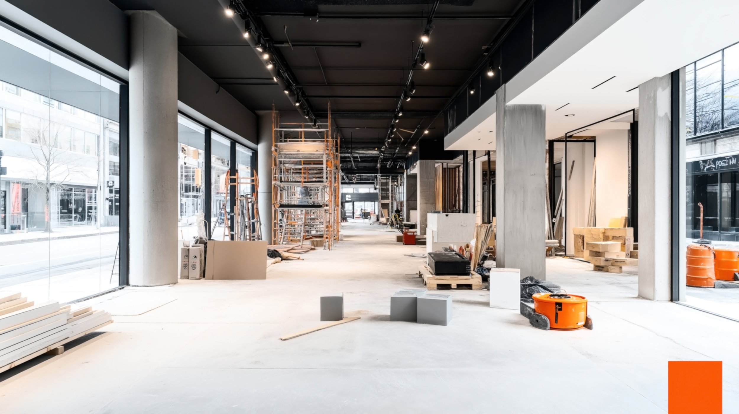 Interior of a commercial space under construction with large windows, scaffolding, construction materials, and tools scattered around.
