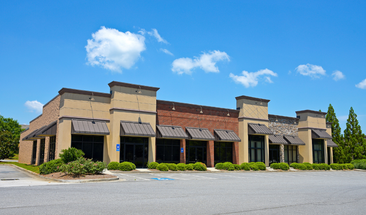 Empty commercial building with large windows, awnings, and a parking lot under a blue sky with clouds.