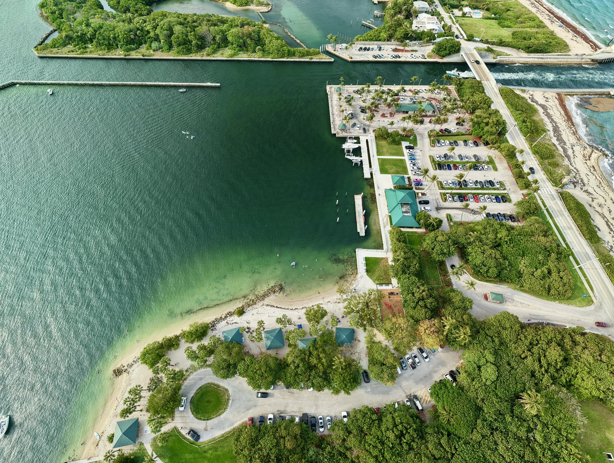 An aerial view of a marina with boats, a parking lot with cars, and a green park area surrounded by water and trees. There are roads with cars and a beach area with sand and waves in the background.