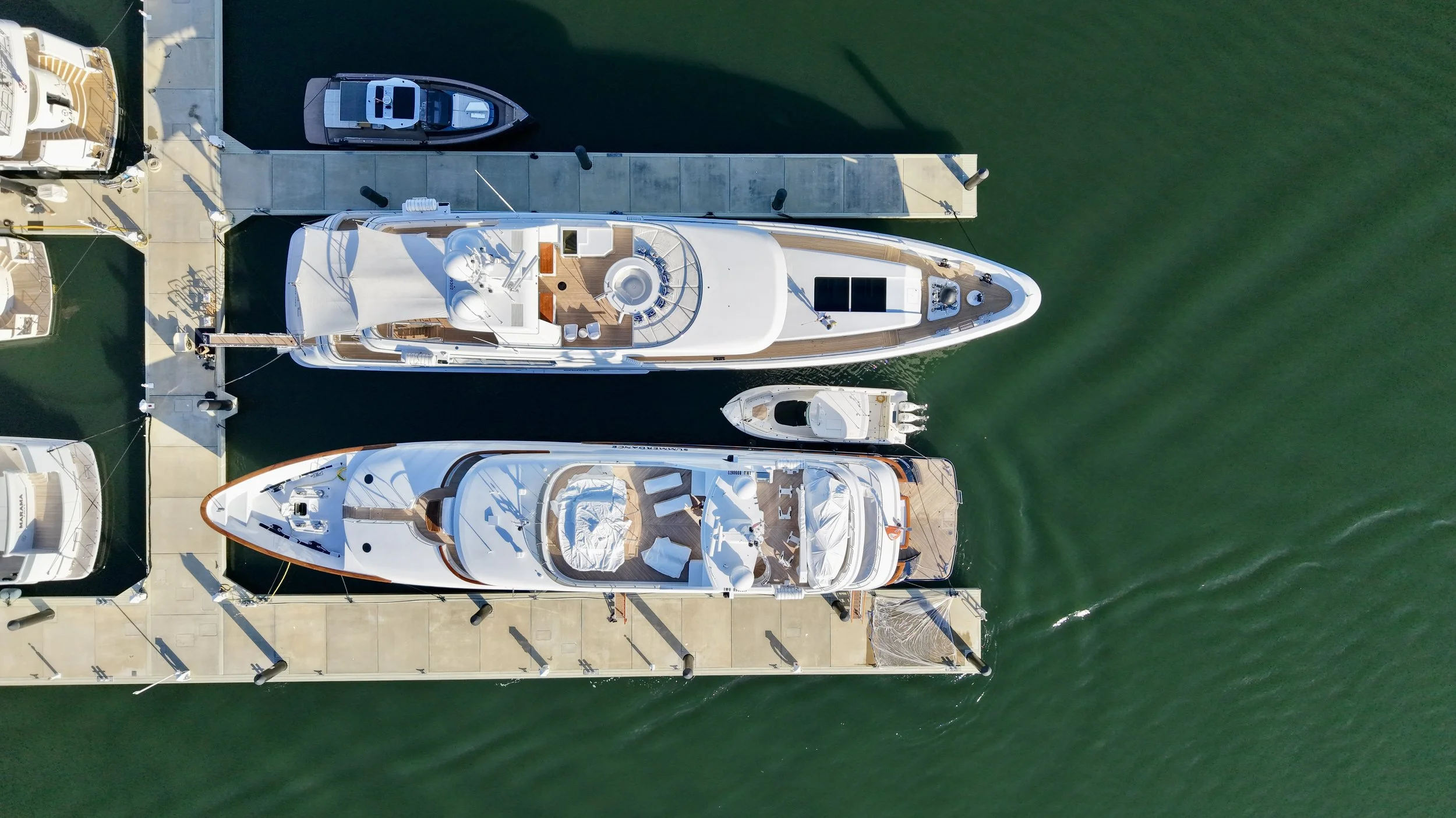 Aerial view of a marina with two large white yachts docked side by side, along with several other boats, all tied to a concrete pier. The water is green, and the boats have decks with seating, sunbeds, and other amenities visible.