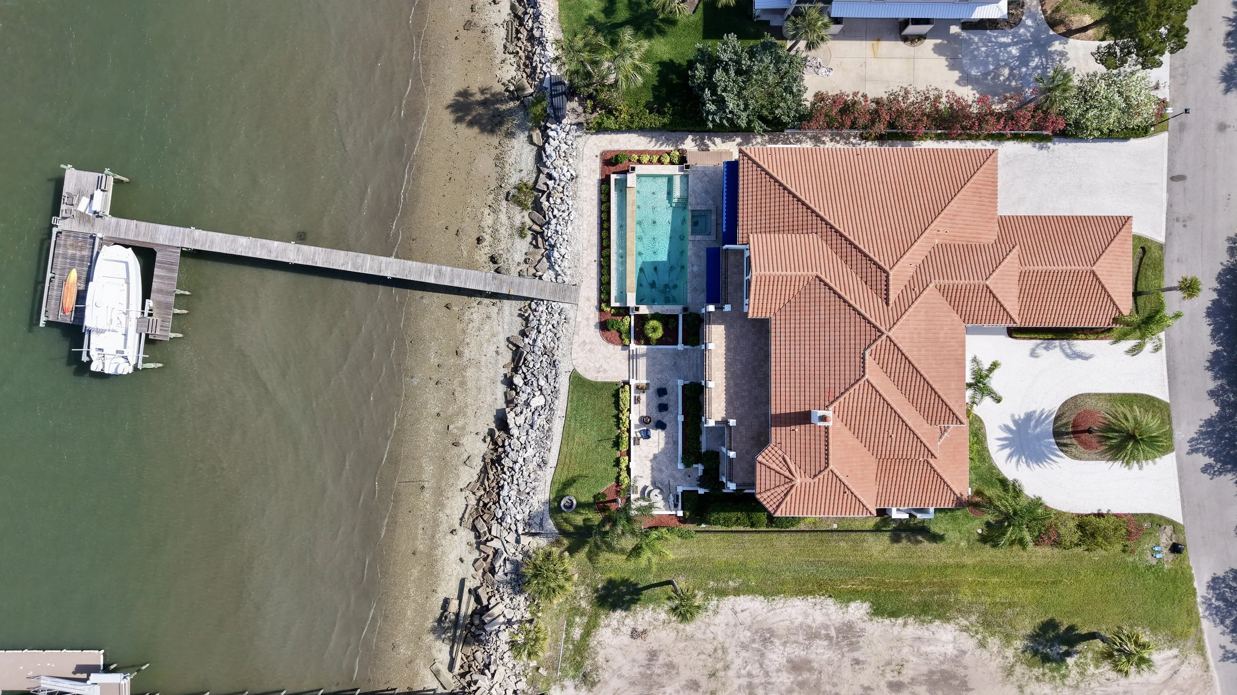 Aerial view of a waterfront house with a swimming pool, landscaped yard, and a dock with a boat.