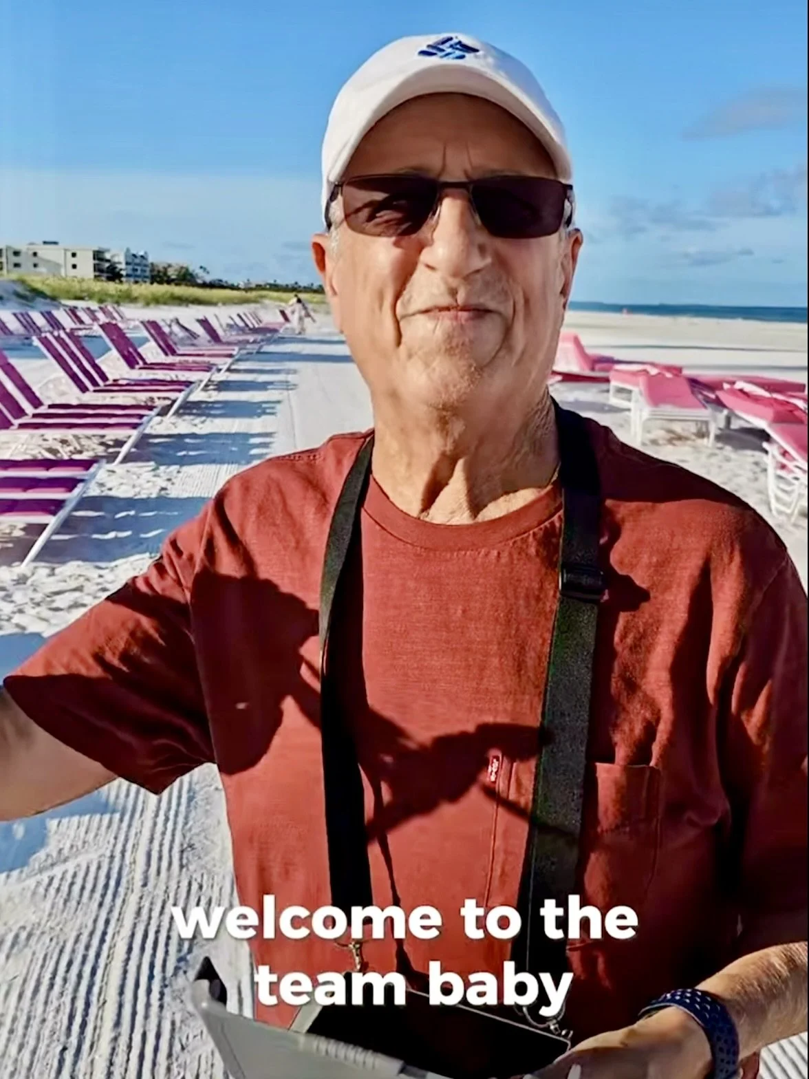 A man wearing sunglasses, a white cap, and a red shirt is being photographed on beach with pink lounge chairs and a sandy shore in the background. The sky is clear and blue, and the man is smiling. Text on the image says "welcome to the team baby."
