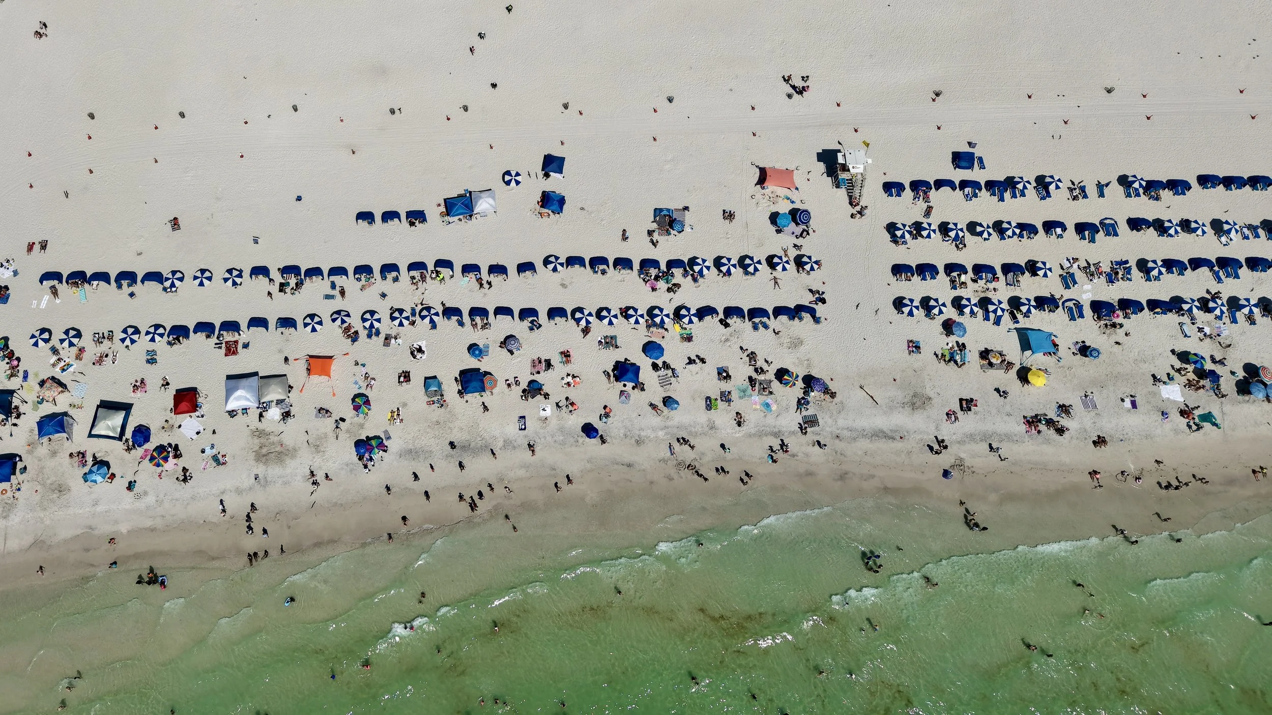 An aerial view of a crowded beach with numerous umbrellas, tents, and people on the sandy shore near the ocean.