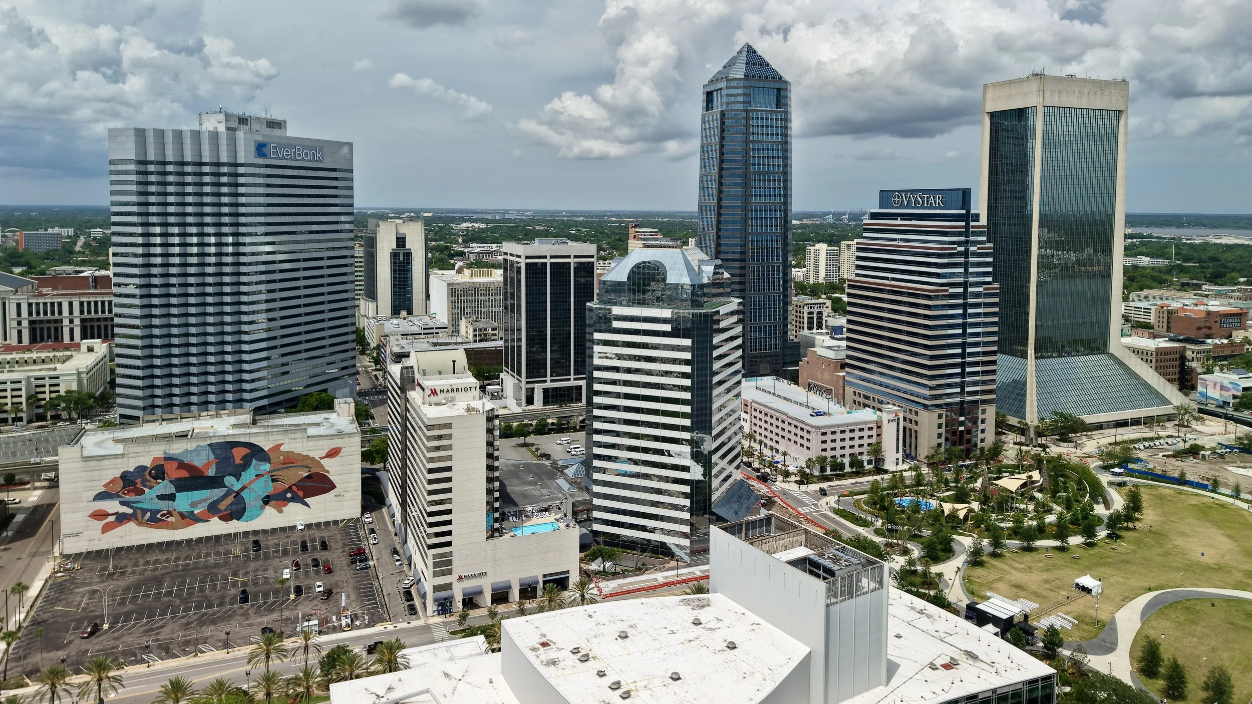 A city skyline with tall modern office buildings, including EverBank and VyStar Tower, under a partly cloudy sky with a park and parking lot in the foreground.