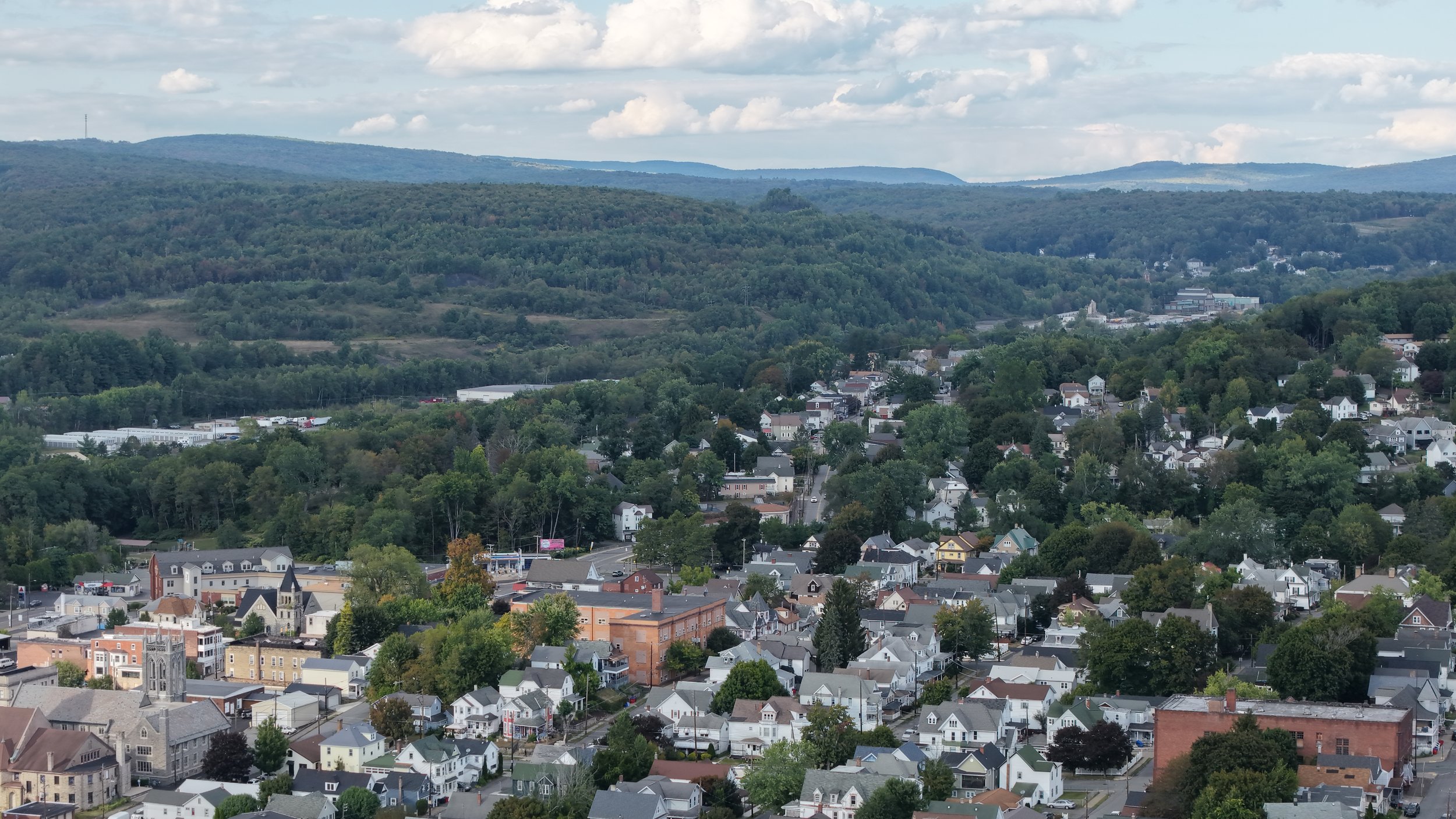 A small city or town with numerous houses, streets, and a church with a tall steeple, surrounded by green trees and rolling hills in the background, under a partly cloudy sky.