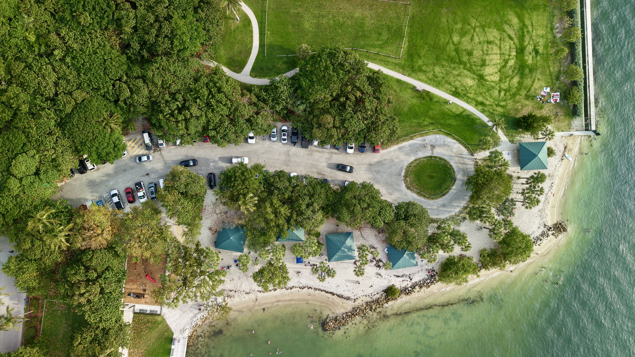 An aerial view of a park near water with green grass, trees, a parking lot, and a small pavilion.