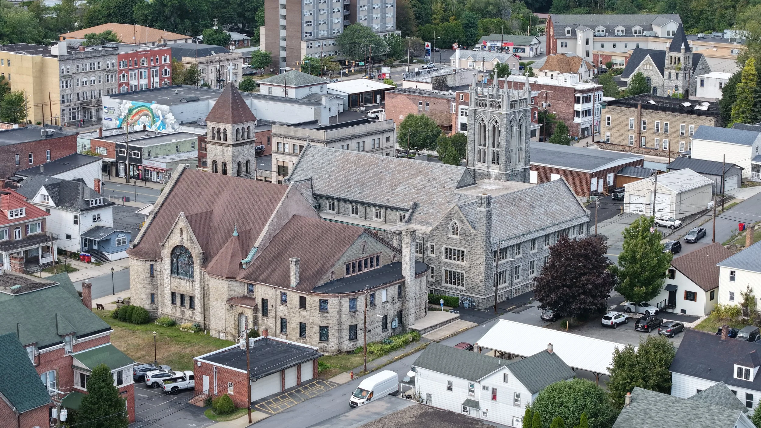 Aerial view of a small town with historic and modern buildings, including churches, houses, and commercial structures, surrounded by trees.