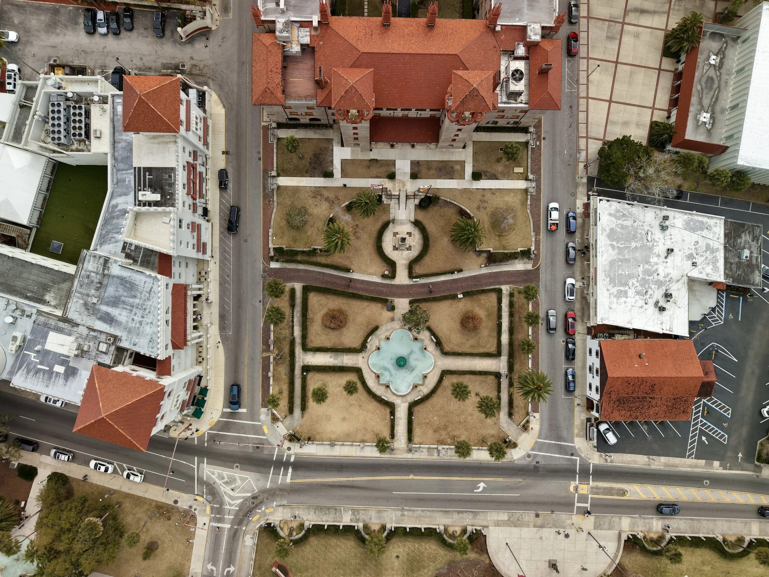 An aerial view of a city block showing a large building with a red-tiled roof, a landscaped courtyard with palm trees, and surrounding streets with parked cars.