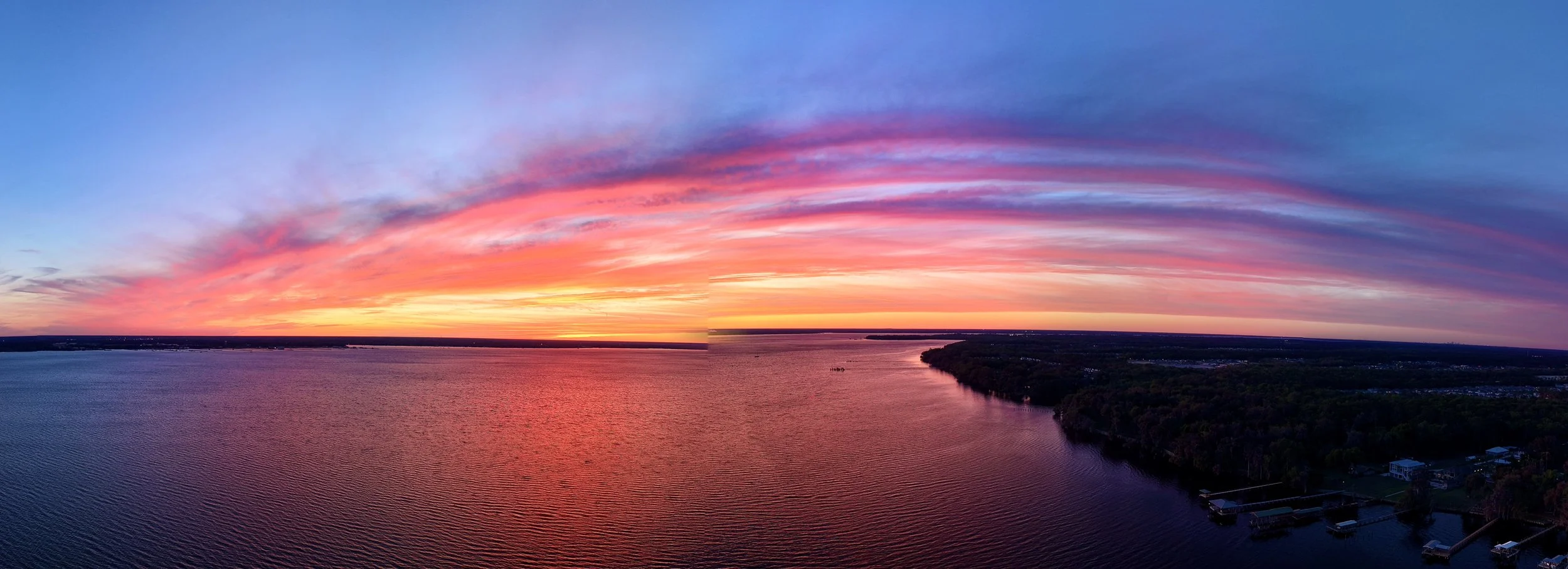 A vibrant sunset over a large body of water with colorful clouds in the sky, and a shoreline with trees and some buildings in the distance.
