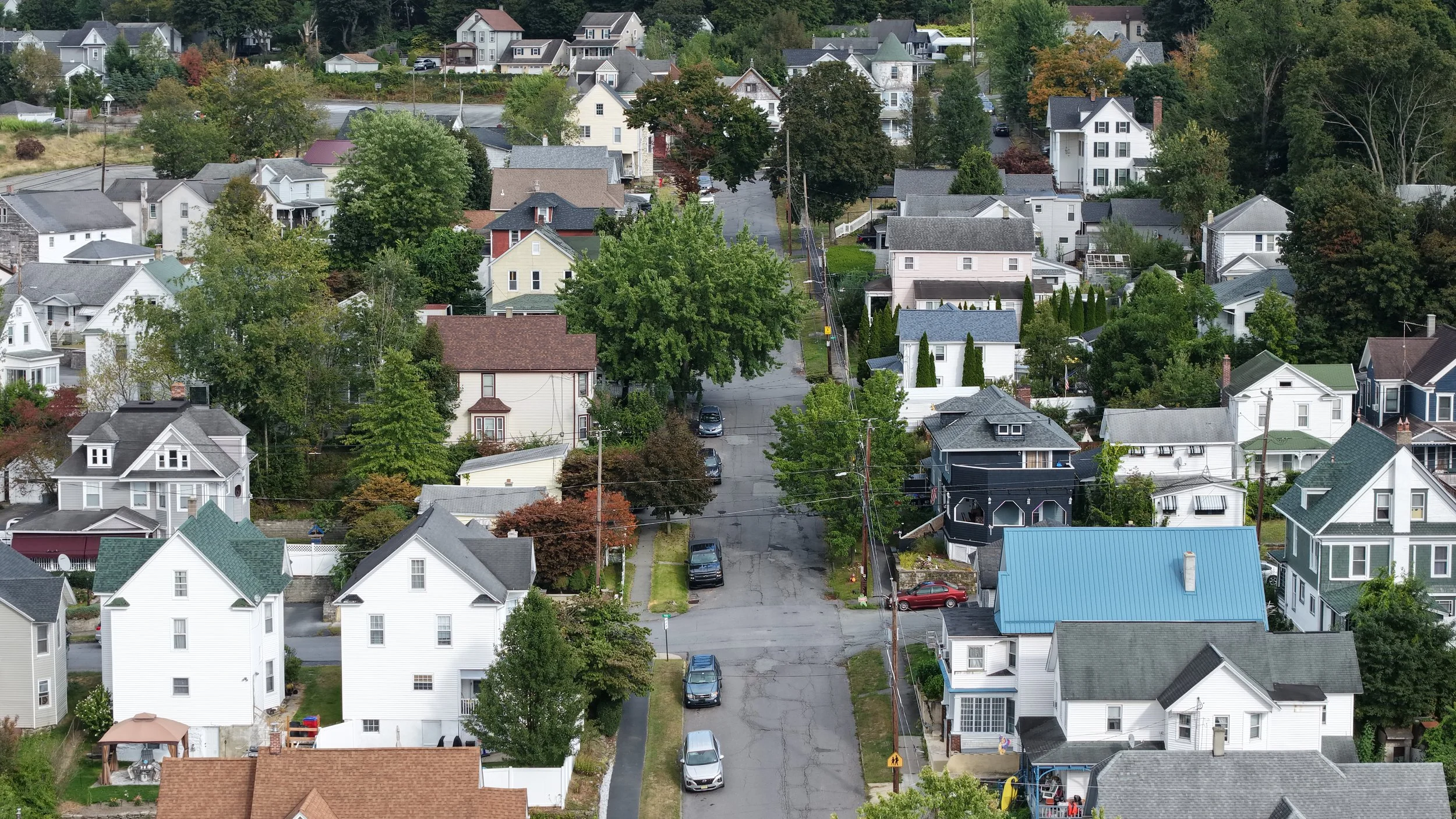 Aerial view of a suburban neighborhood with multiple houses, trees, and parked cars along a street.