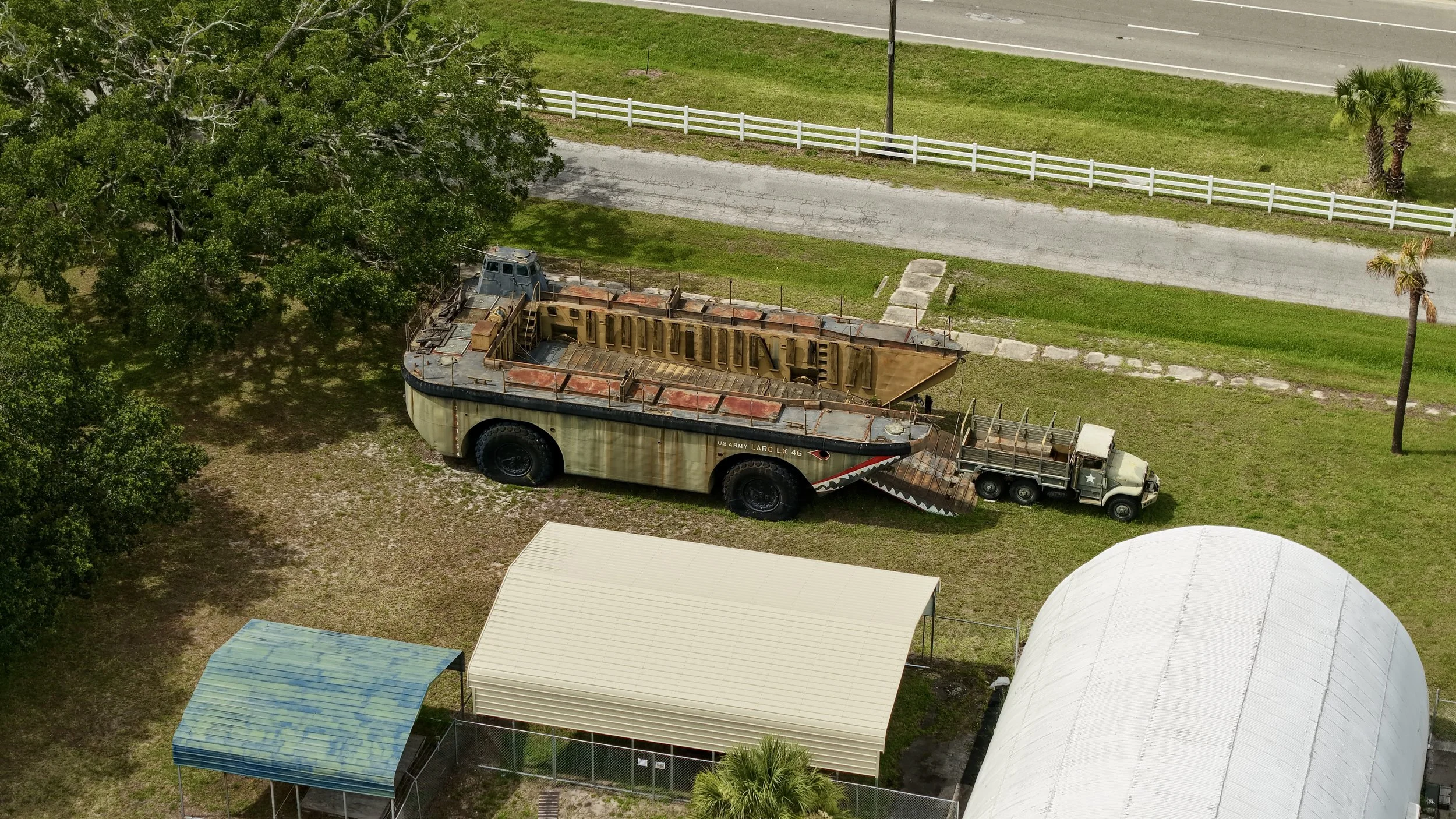 An aerial view of a large military amphibious vehicle parked near a white picket fence, with a truck attached to it. The scene includes trees, a grassy area, sidewalks, and some small buildings or structures.