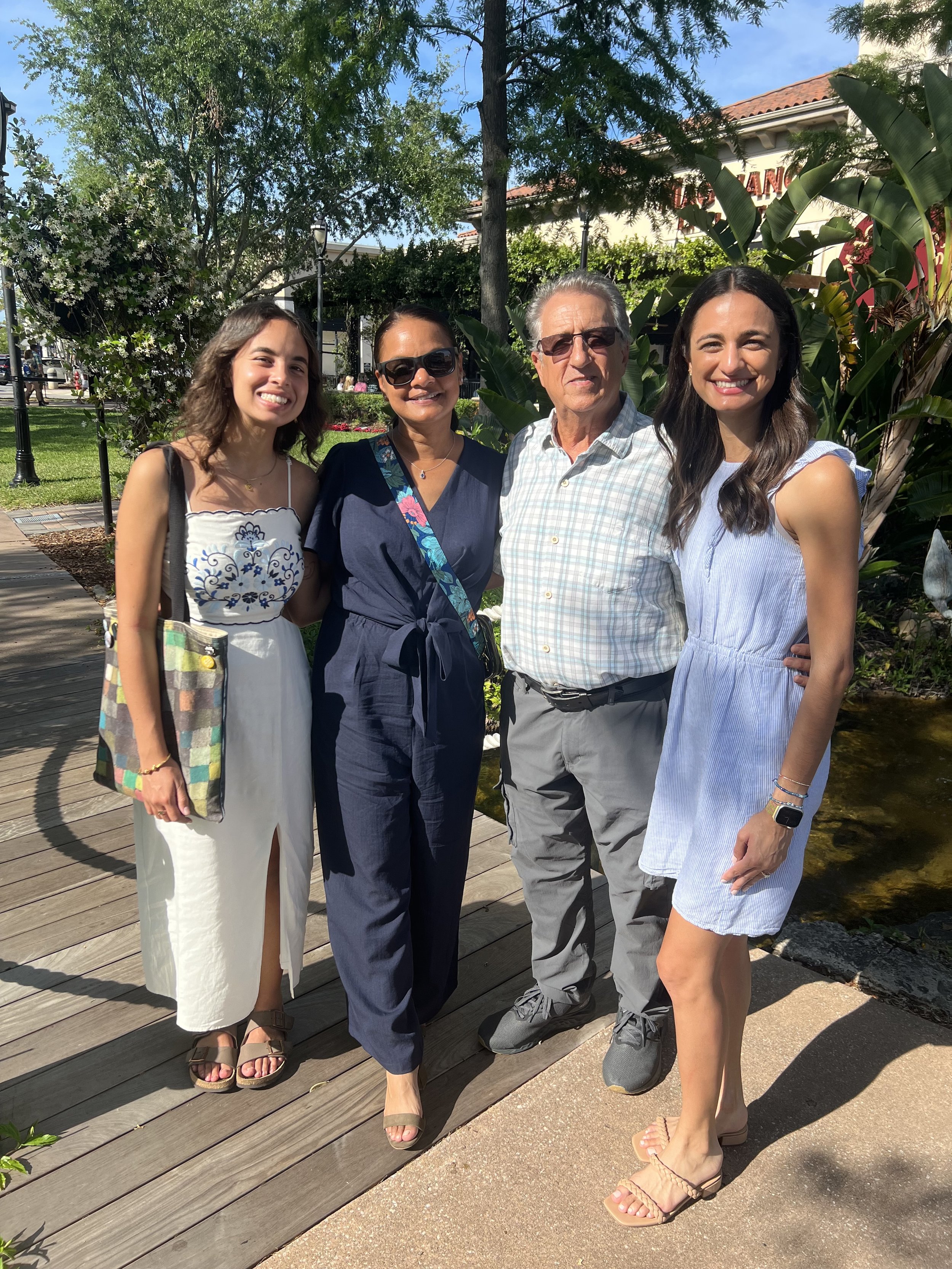 A family of four standing outdoors on a sunny day, smiling, with trees and a building in the background.