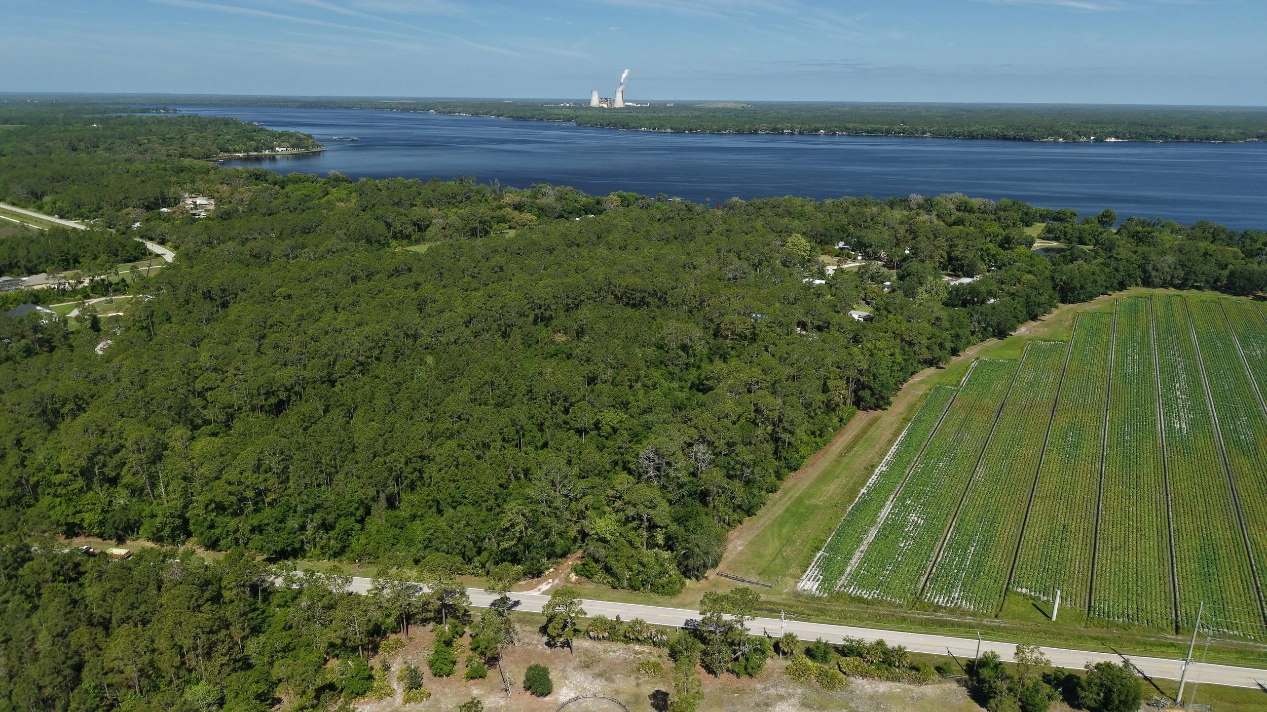 An aerial view of a large river with dense forested areas on both sides, some roads, and green agricultural fields in the foreground. In the distance, there are industrial cooling towers emitting steam or smoke.