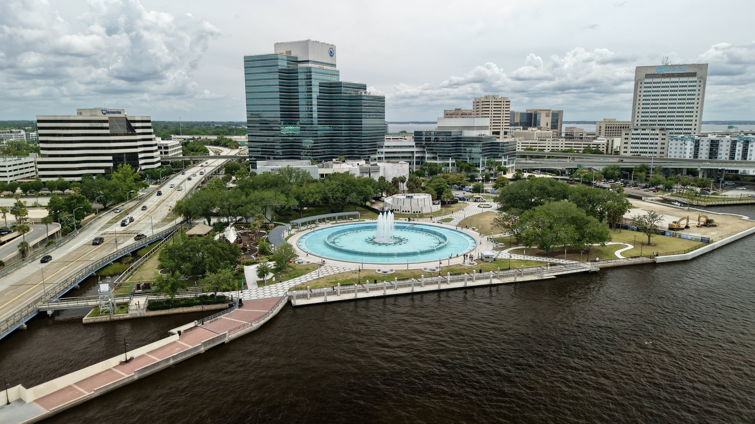 A city skyline with tall modern buildings, a large circular fountain with a water jet in the front, and a river running alongside a park area with trees and pathways.