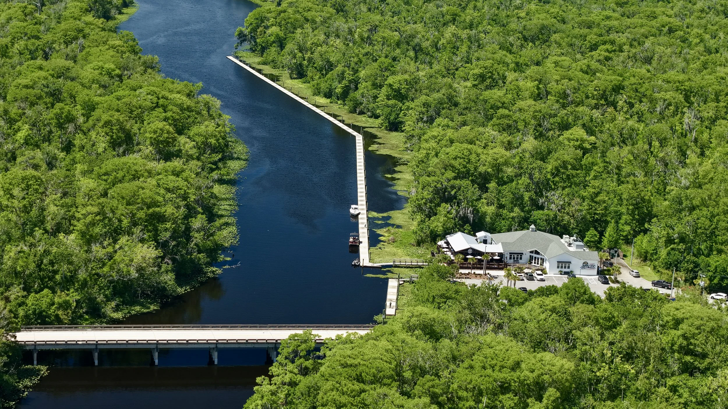 Aerial view of a marina with boats docked along a waterway, surrounded by dense green trees, and a building with a parking lot nearby.