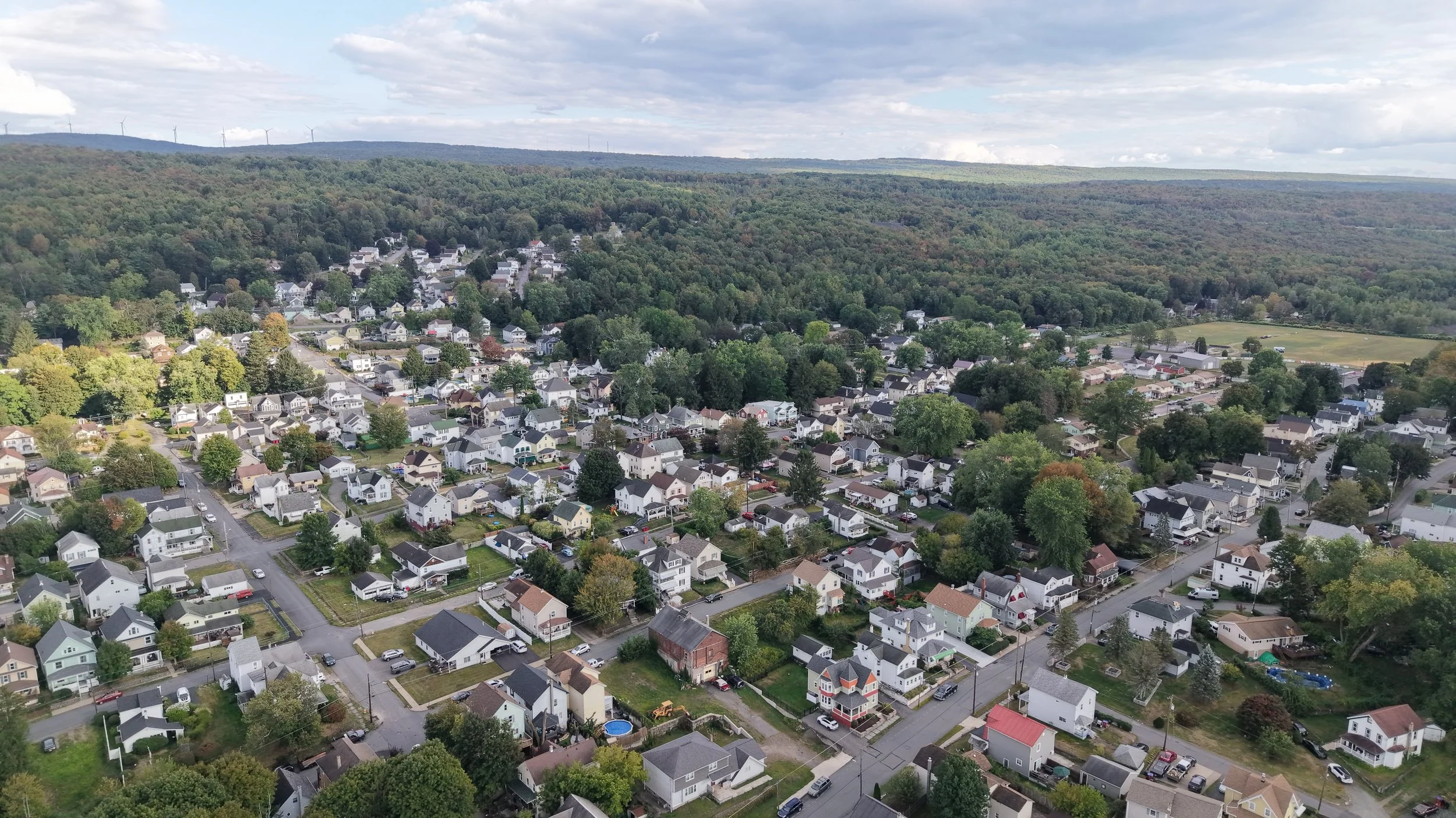 An aerial view of a suburban neighborhood surrounded by a lush, green forest, with many houses, streets, and some open spaces.