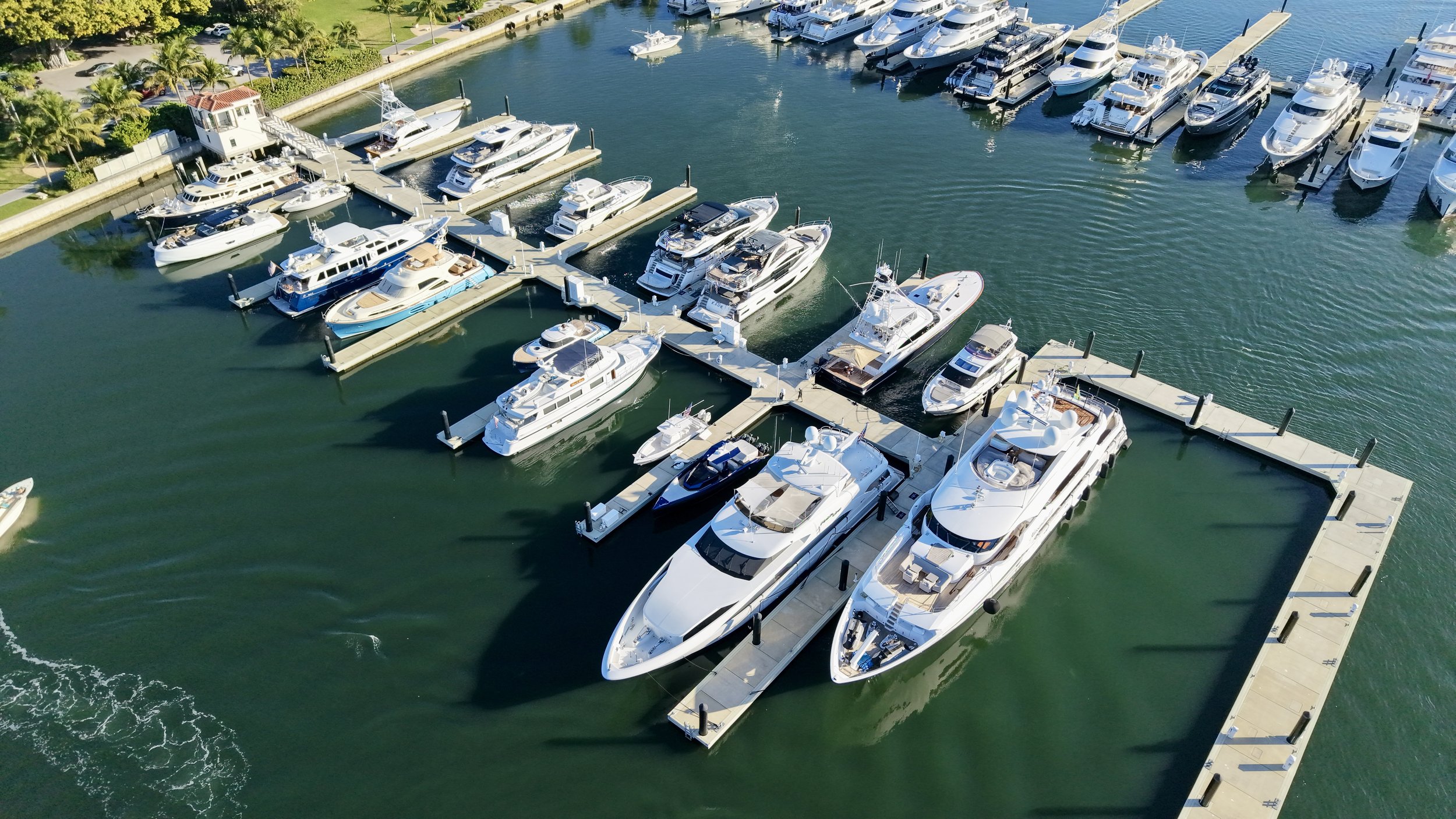 Aerial view of a marina with multiple yachts and boats docked along floating piers, surrounded by water and a nearby green park area with palm trees.