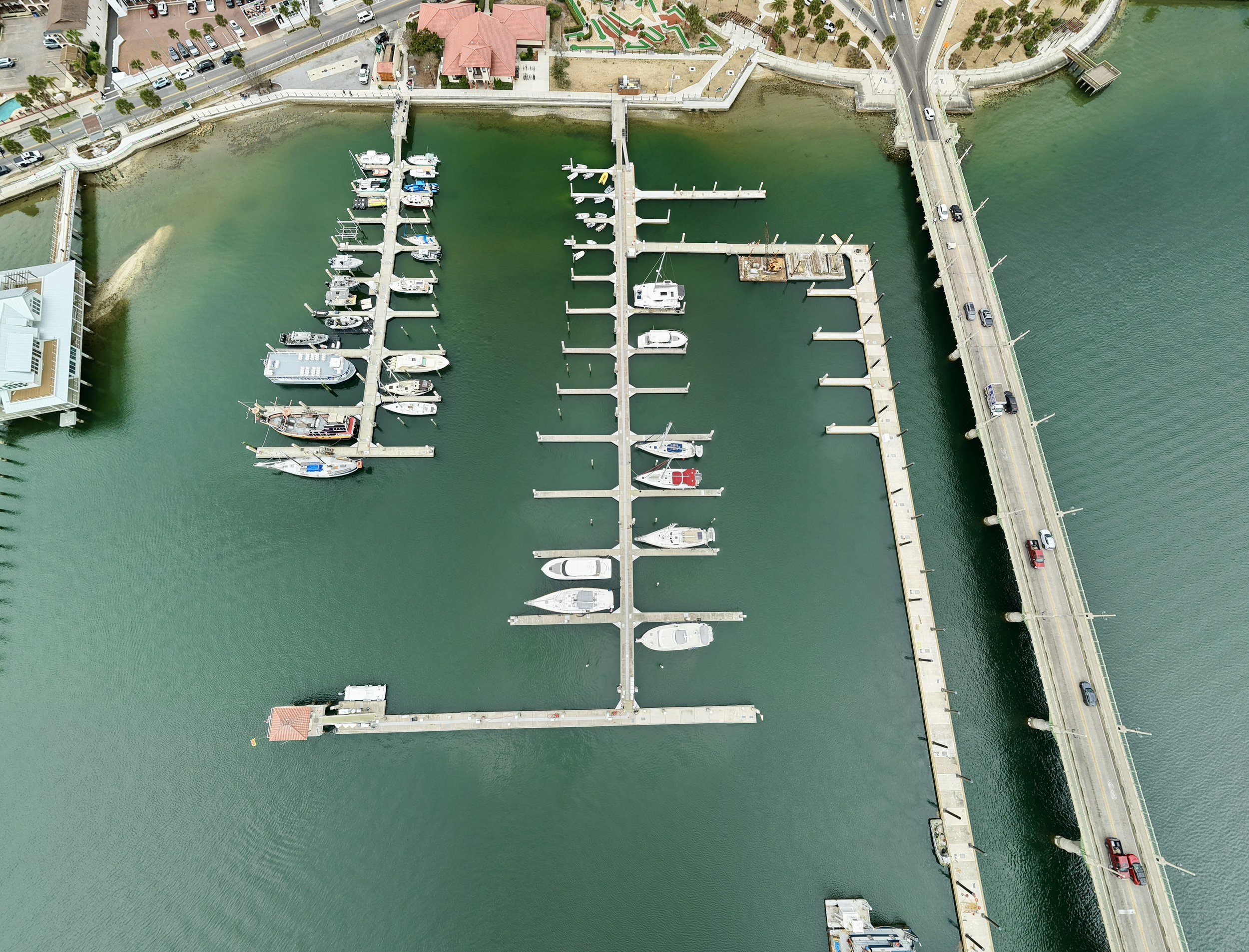 An aerial view of a marina with boats docked at piers, surrounded by a cityscape including roads, buildings, and a park area with pathways and trees.