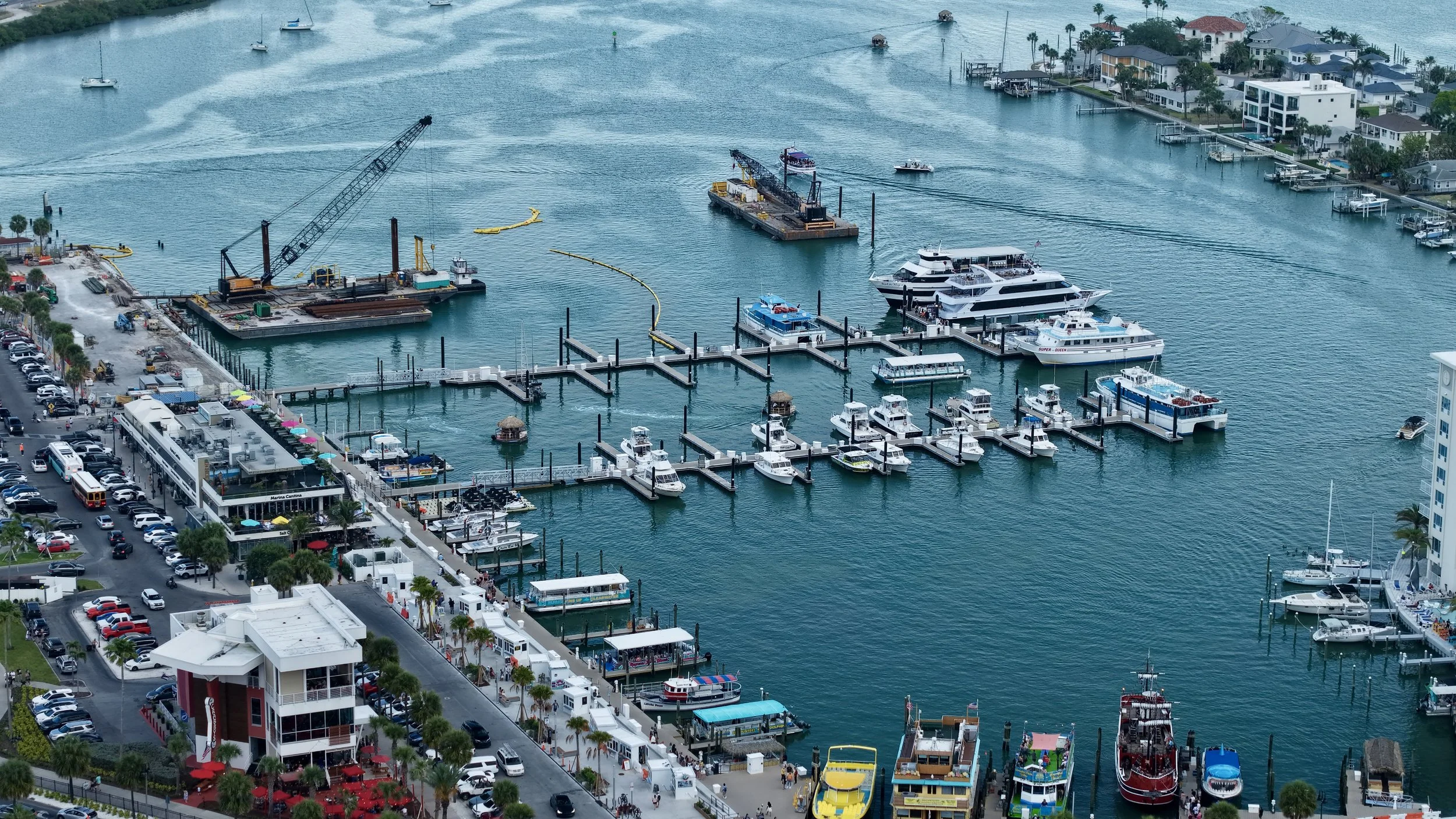 Aerial view of a marina with numerous boats and yachts docked, a large construction platform with cranes, a busy parking lot with cars, a building with outdoor seating, and a waterfront neighborhood with houses and palm trees.