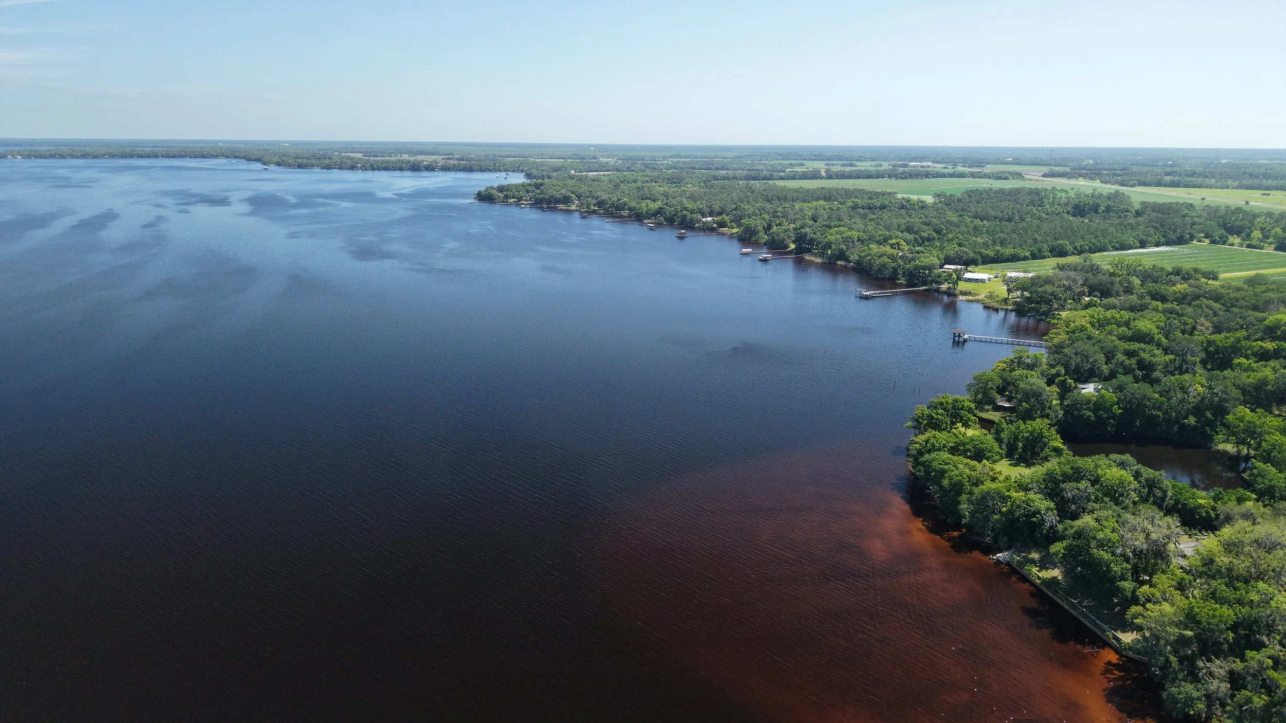 Aerial view of a large body of water bordered by a lush green landscape with trees and open fields, some docks and structures along the shore, and a clear sky overhead.