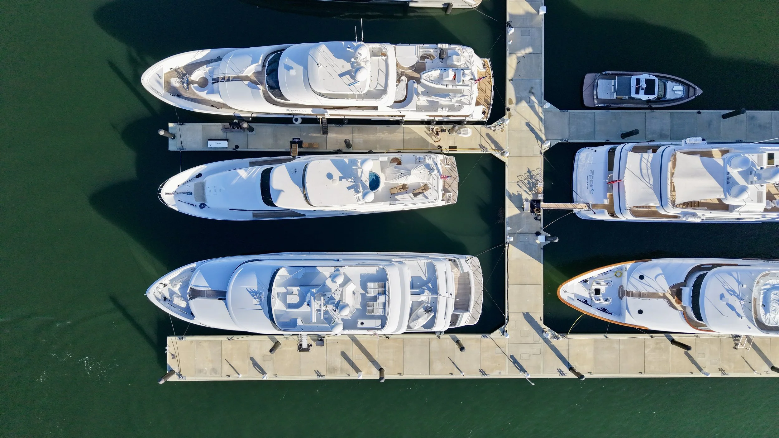 Aerial view of a marina with multiple yachts docked along the pier, casting their shadows on the water.