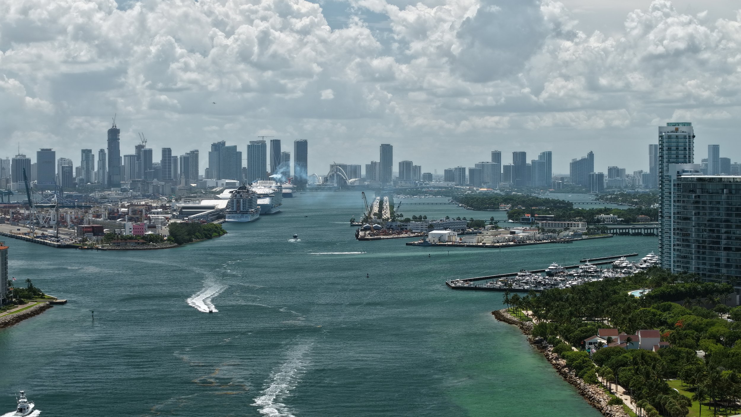Wide view of a city skyline with tall buildings and skyscrapers, a waterway with boats and yachts, and a marina with piers, under a partly cloudy sky.