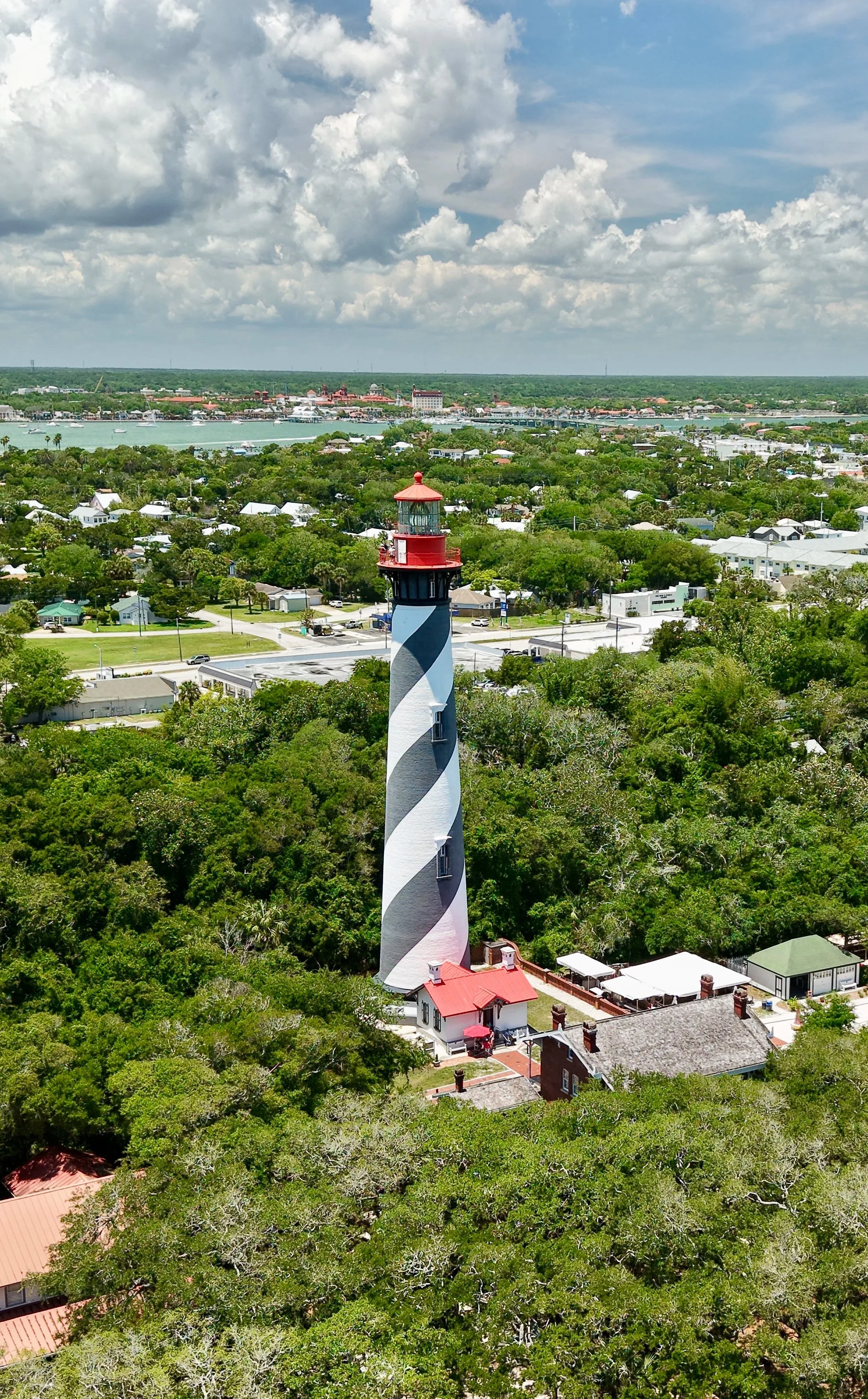 St. Augustine Lighthouse and Maritime Museum with black and white spiral stripes and a red top, surrounded by green trees and residential houses in a Anastasia Island, Florida, , with water, boats, and a cloudy sky in the background.