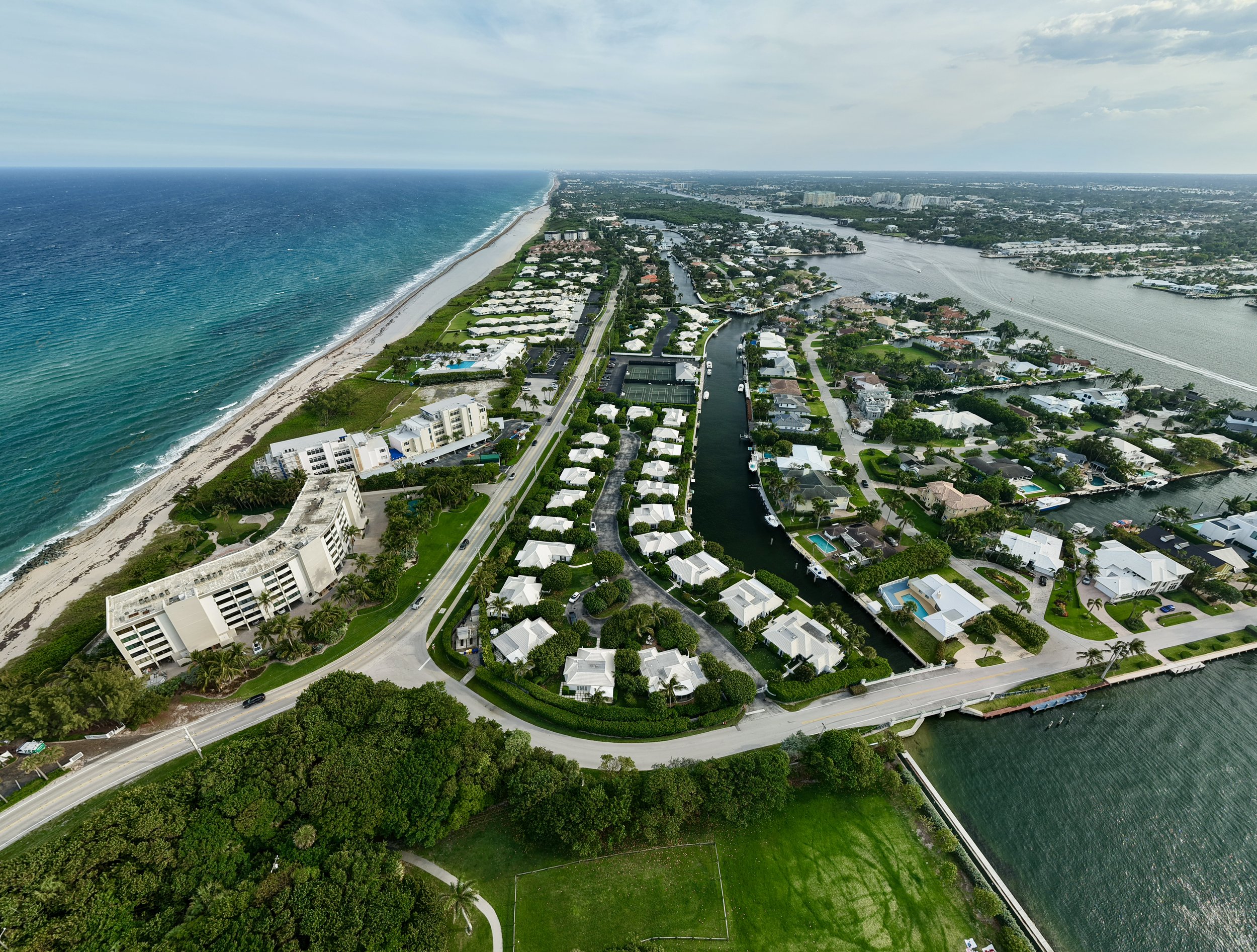 An aerial view of a coastal town with white buildings, a beach, and a canal system. The ocean is on the left, and the town extends inland with residential and commercial areas.