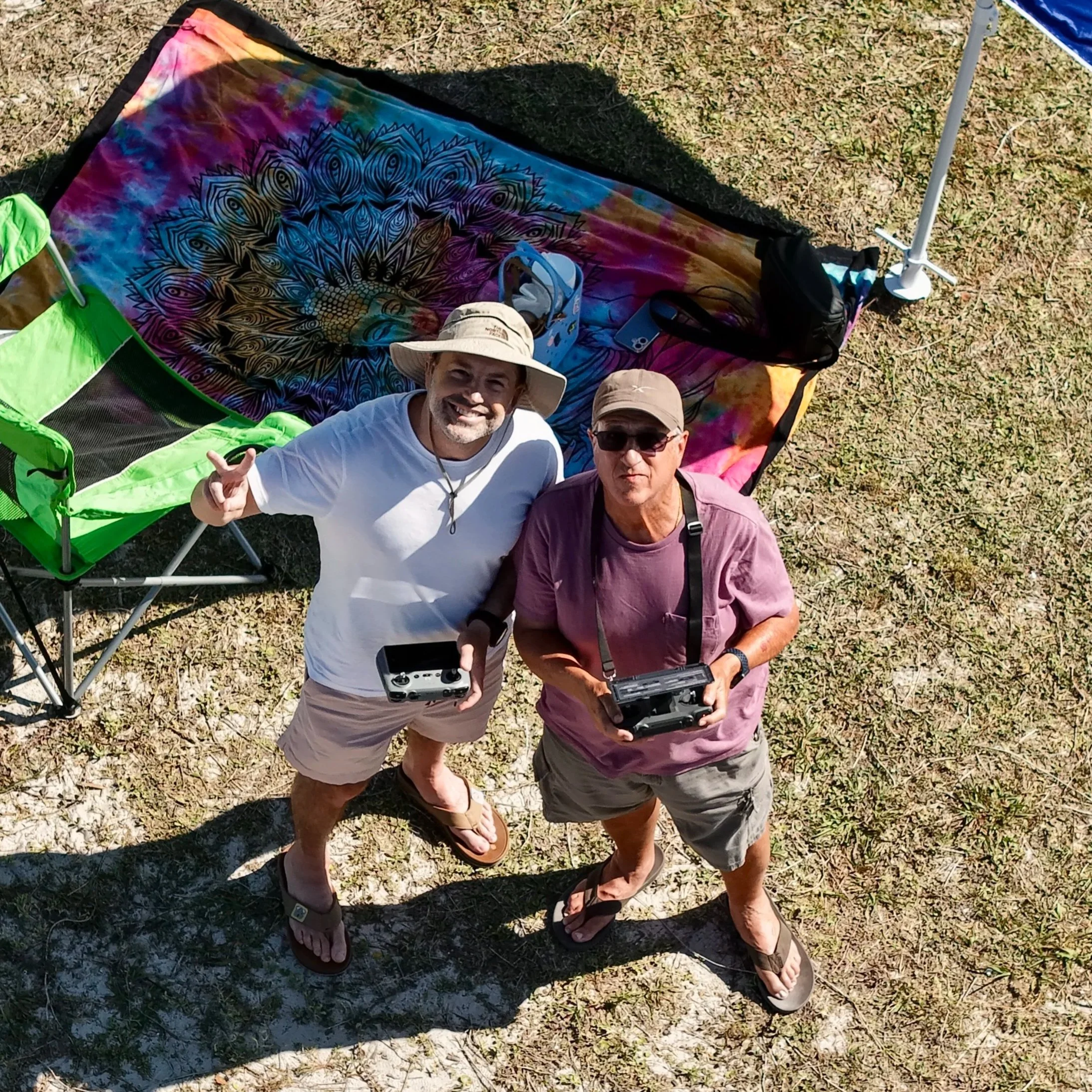 Two men in casual clothes and hats standing on grass, taking a selfie with a drone in a park or outdoor area. The photo is captured from above, showing a colorful tie-dye blanket and some equipment around them.
