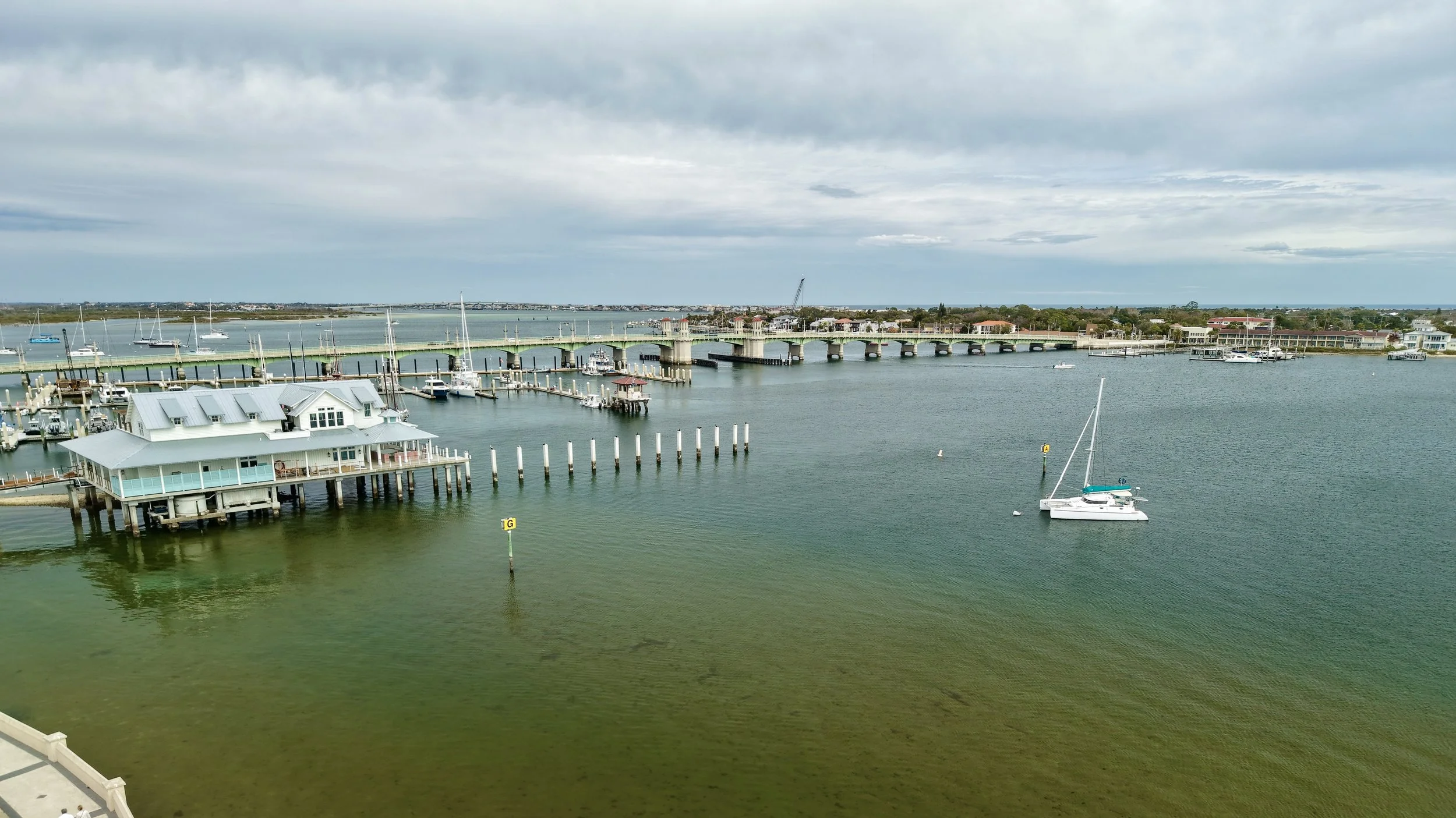 A marina with several sailboats and yachts docked, a bridge in the background, buildings along the shoreline, and a cloudy sky overhead.
