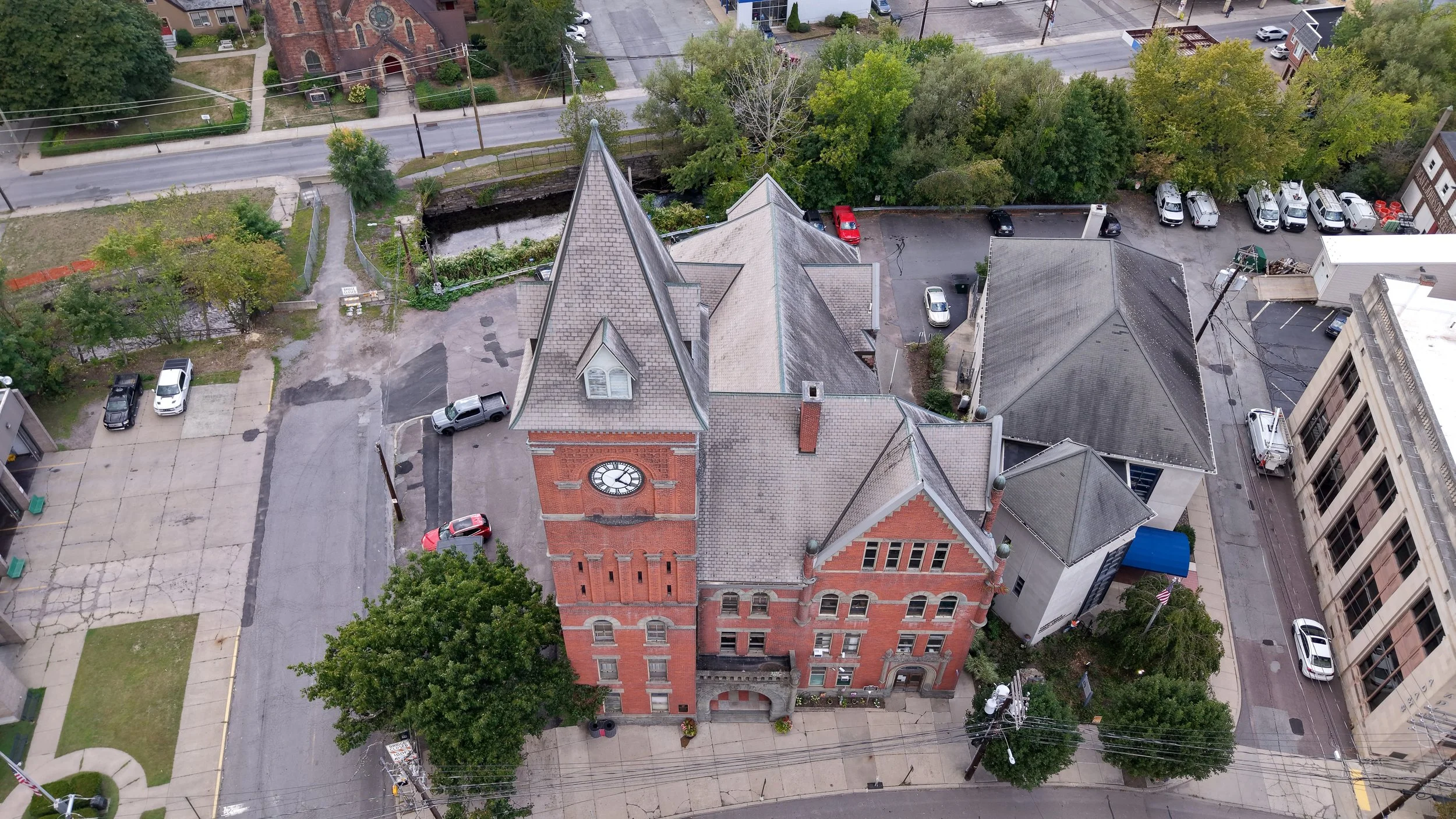 Aerial view of a historic red brick church with a clock tower, surrounded by trees, parking lot, and city streets, with residential and commercial buildings nearby.