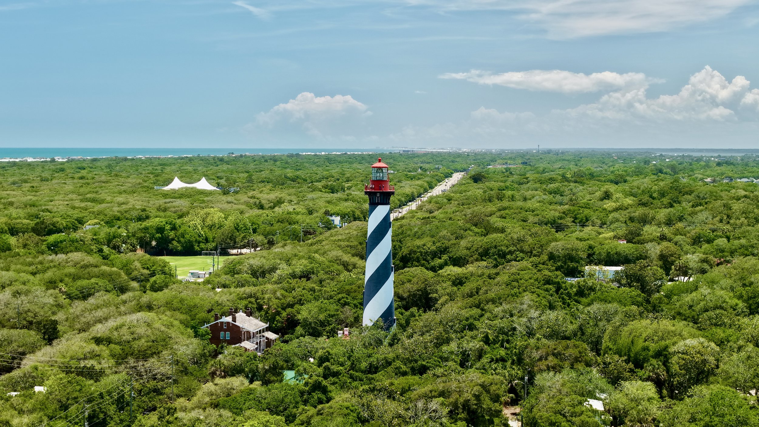 A coastal landscape with a striped lighthouse in the foreground, surrounded by green trees and a road leading towards the horizon, with a view of the ocean and partly cloudy sky in the background.