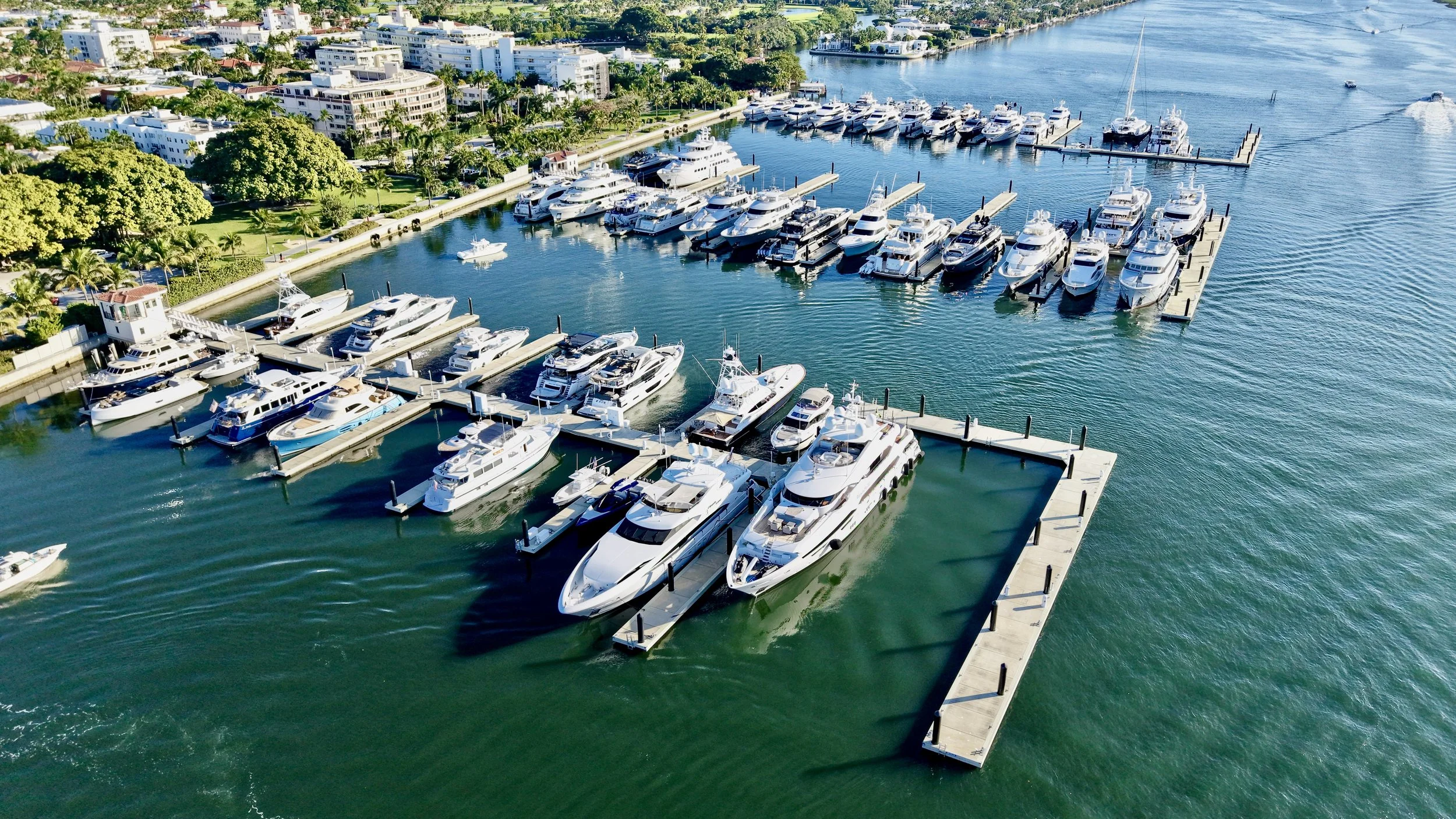 An aerial view of a marina filled with numerous white yachts and boats docked along the piers, with a backdrop of a cityscape and waterway.