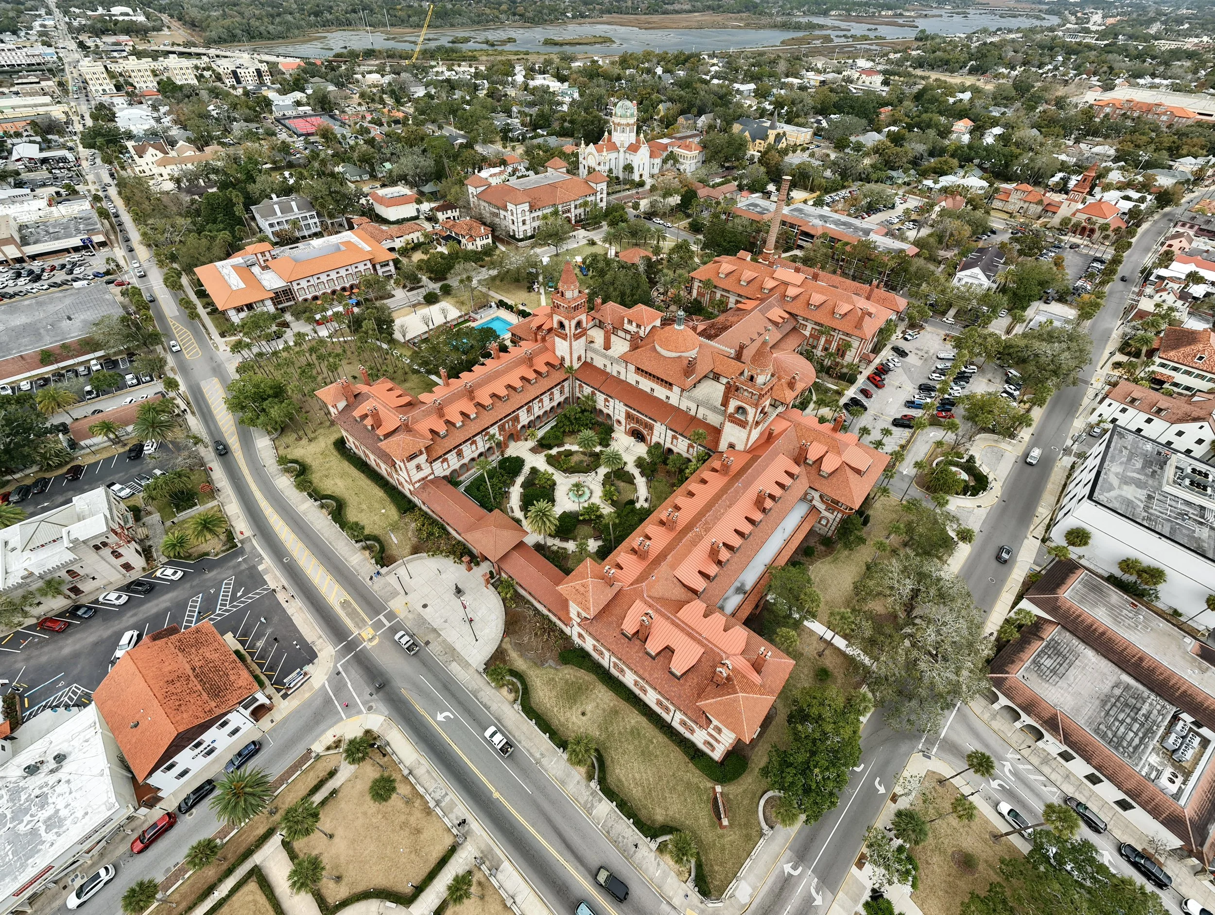 Aerial view of a city with a mix of historic and modern buildings with red-tiled roofs, a park with a pool, streets, and parking lots.