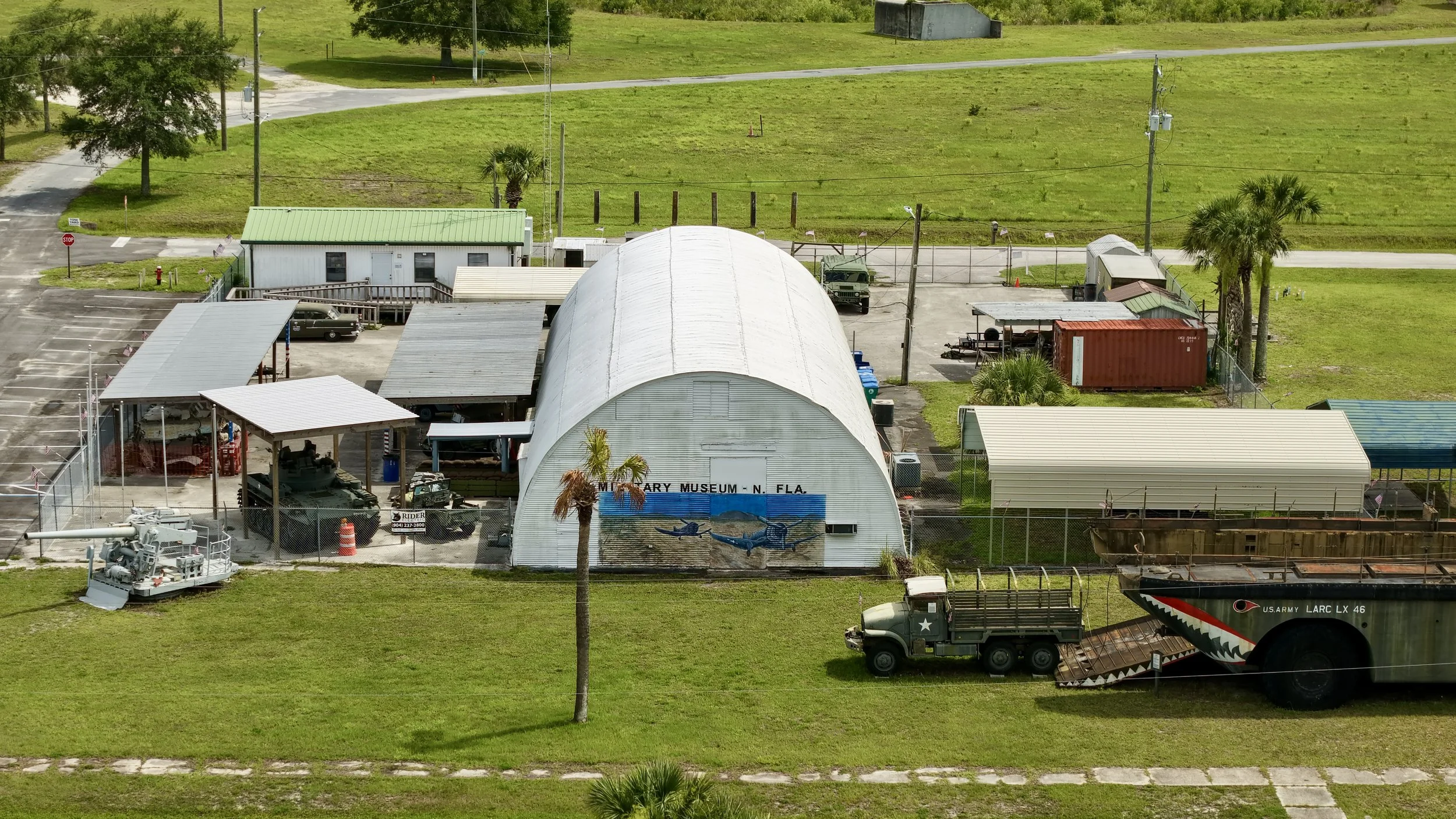 Aerial view of a military museum lot with various military vehicles, a white hangar with painted whales and the text 'Military Museum - N. Fla.', smaller sheds, and palm trees, enclosed by chain-link fencing, with open grassy areas and a road in the 