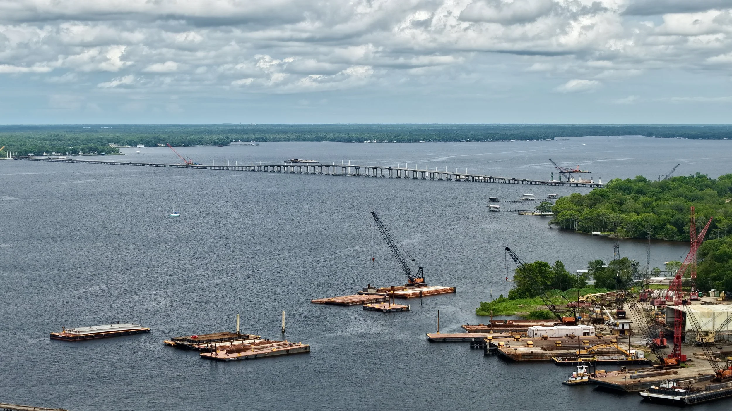 View of a large river with a bridge under construction, multiple cranes, floating platforms, and green tree-covered areas.
