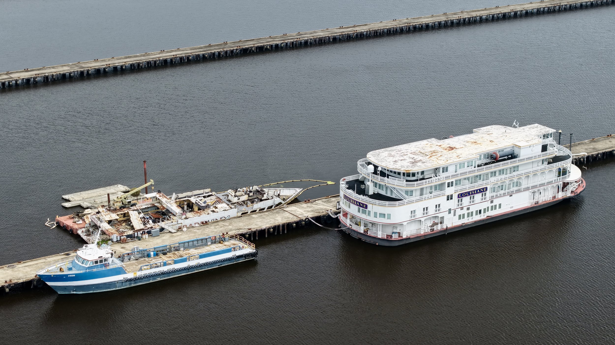 Aerial view of a harbor with two boats docked at a pier. One boat is a large white multi-deck vessel named 'Louisiane,' and the other is a smaller blue and white vessel named 'Yision.' A section of the pier shows signs of damage or construction, with