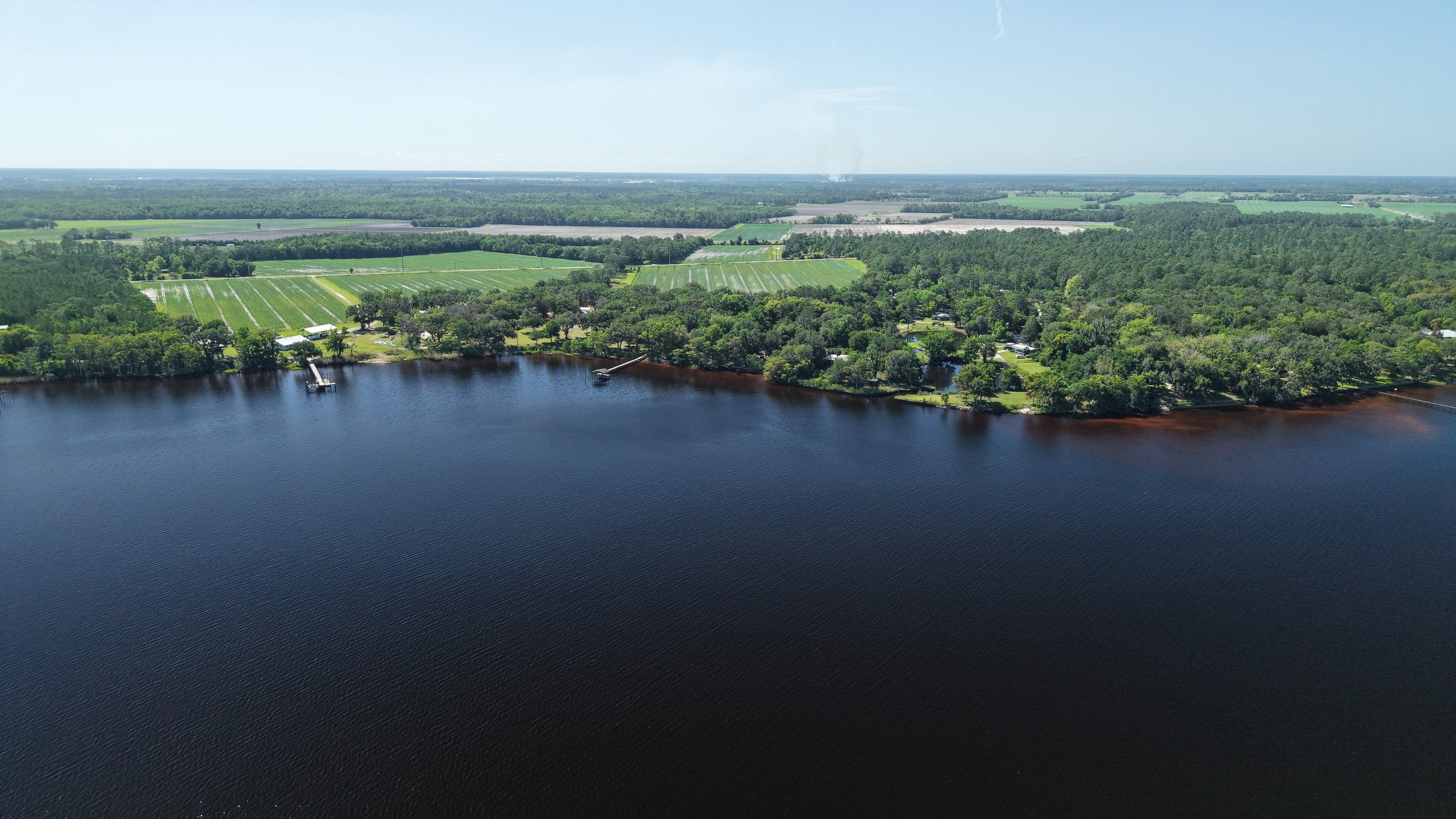 Aerial view of a landscape with a body of water in the foreground and lush green fields and forests in the background.