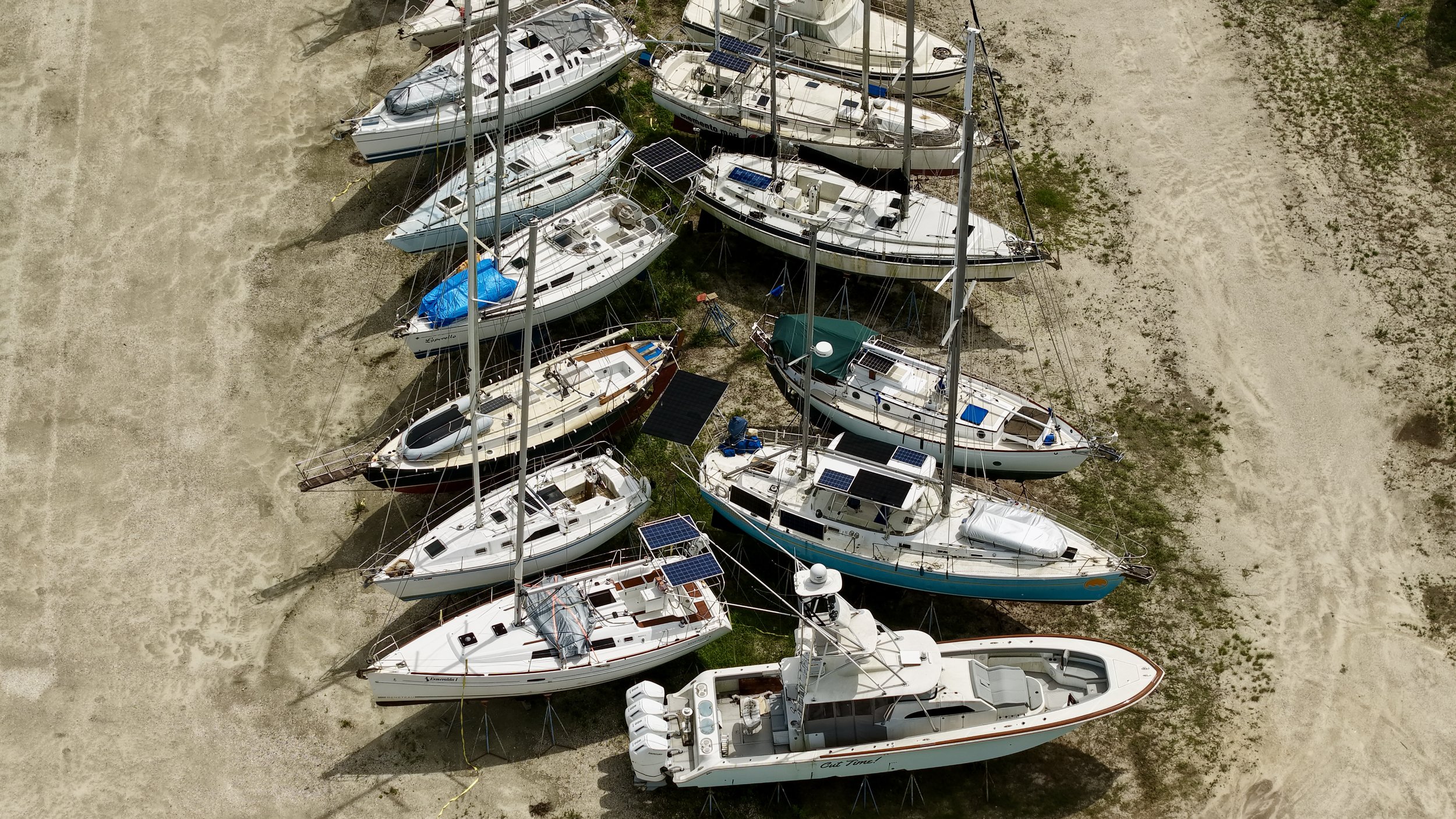An aerial view of multiple sailboats and motorboats docked on a sandy area, some with solar panels on their roofs.