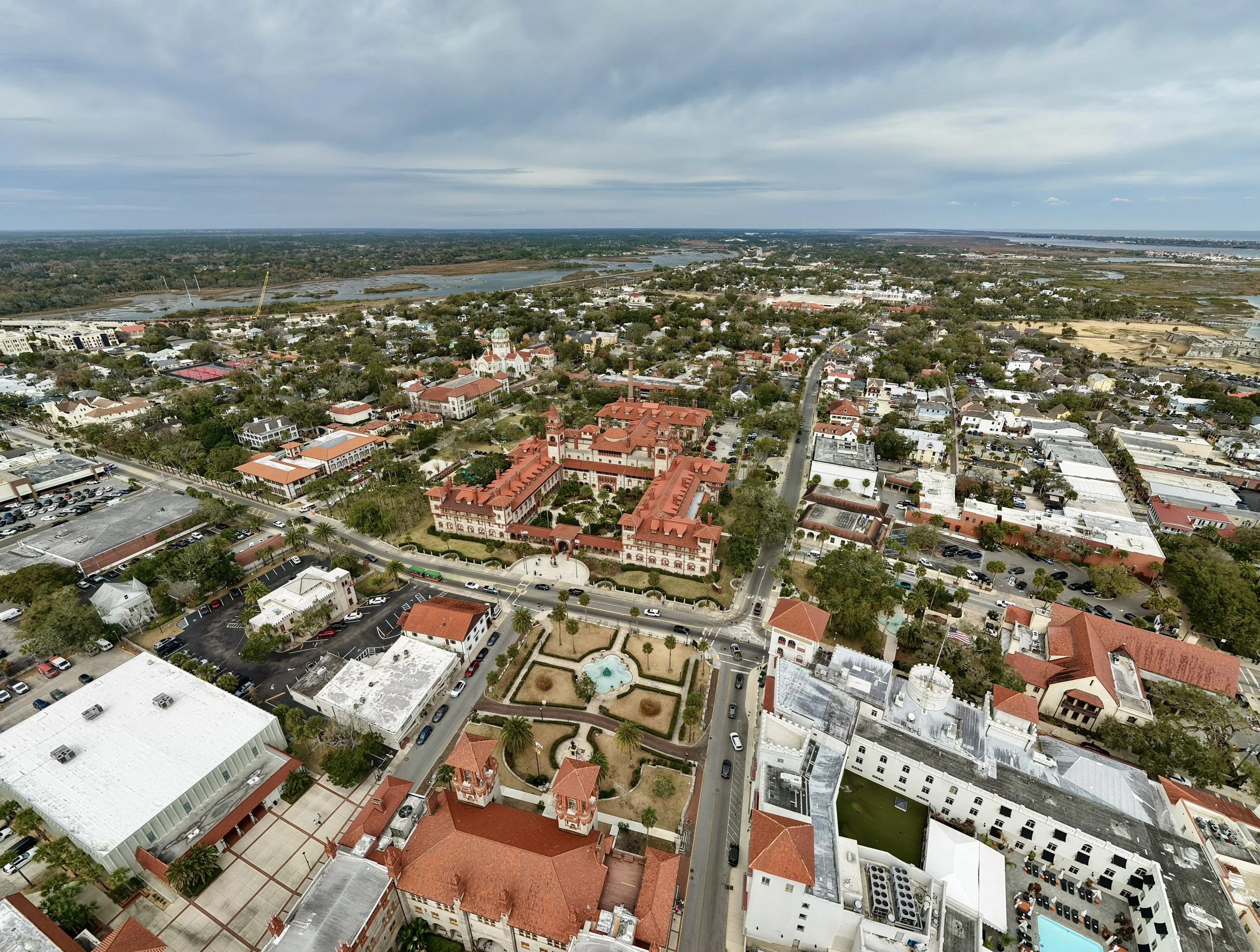 An aerial view of a city with a mix of historic and modern buildings, red-tiled roofs, and streets lined with trees, with a river and green landscape in the background, under a cloudy sky.
