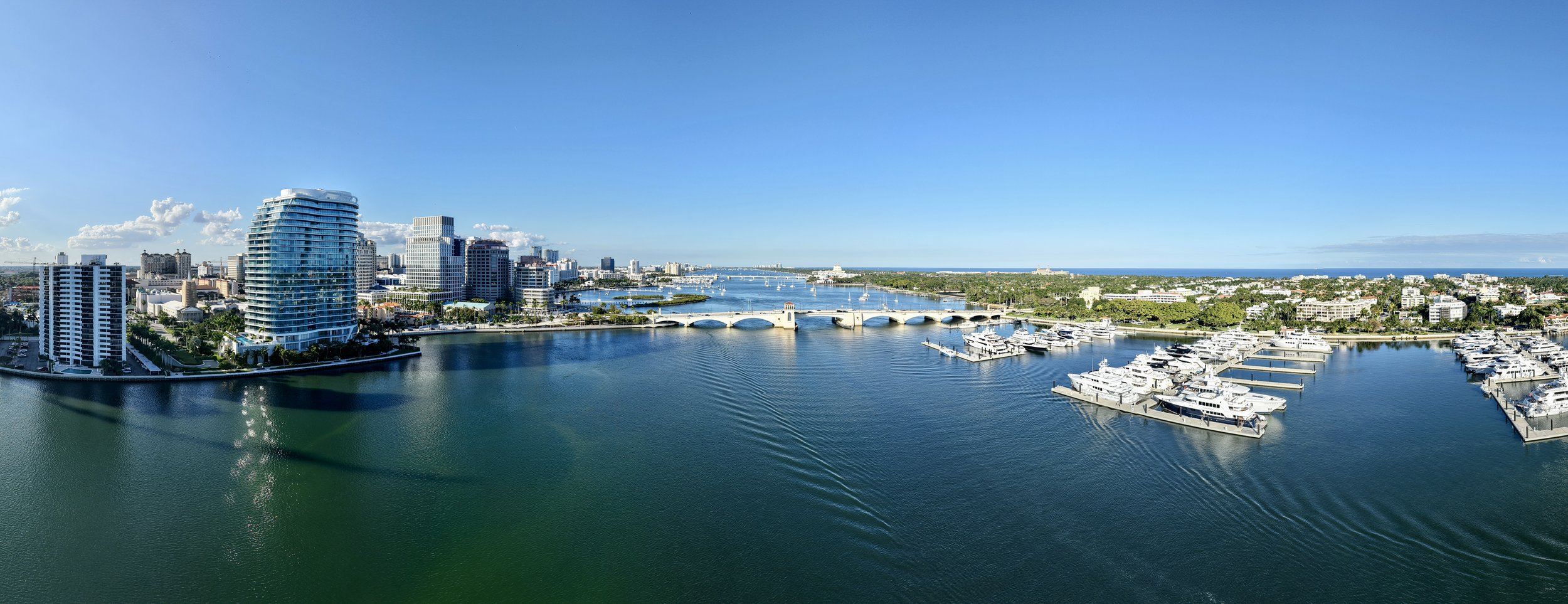 Aerial view of a city skyline by a marina with yachts and boats, high-rise buildings, a bridge over the water, and a clear blue sky.