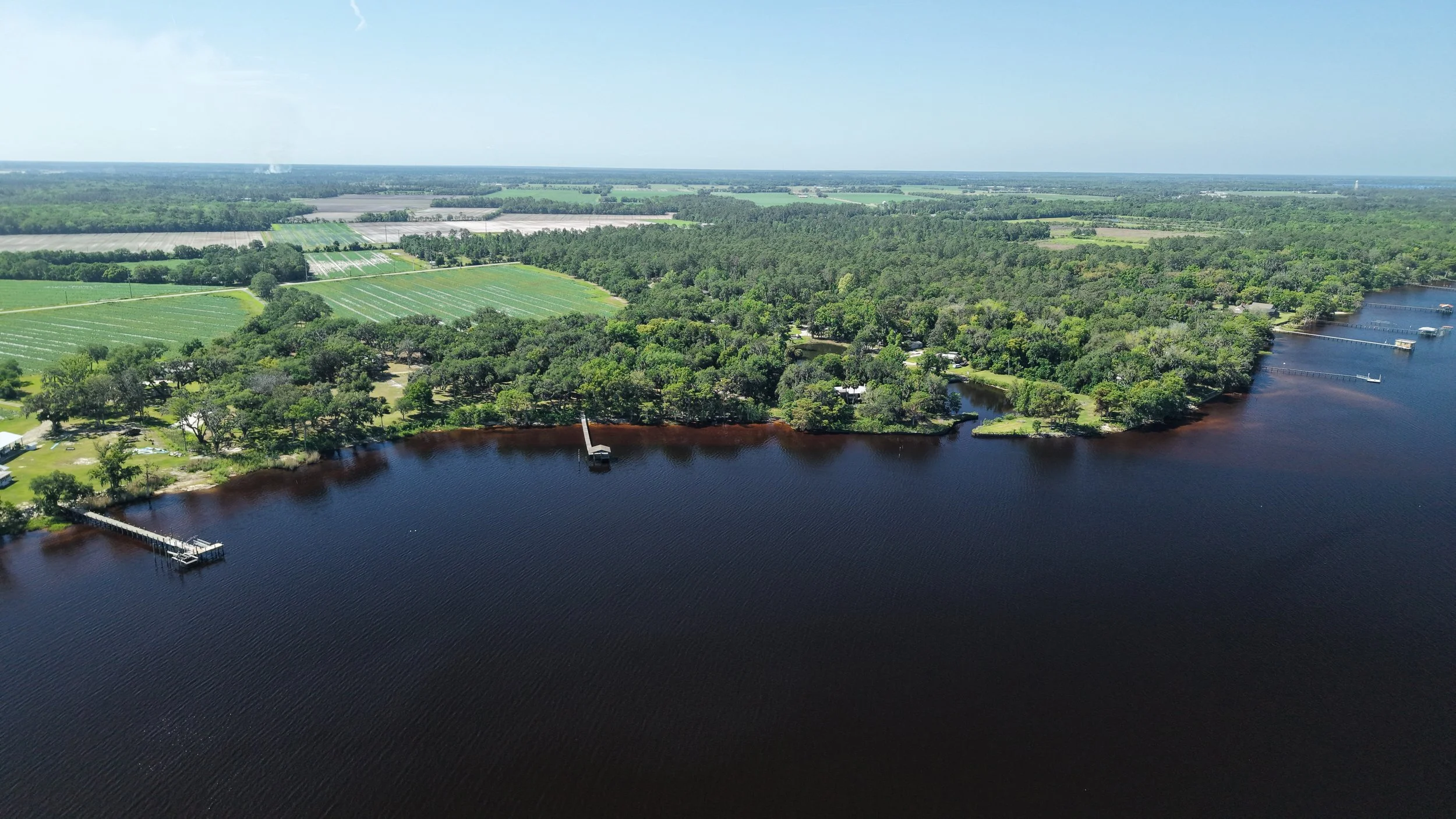 Aerial view of a river with wooded shoreline, boat docks, and surrounding farmland and forest.