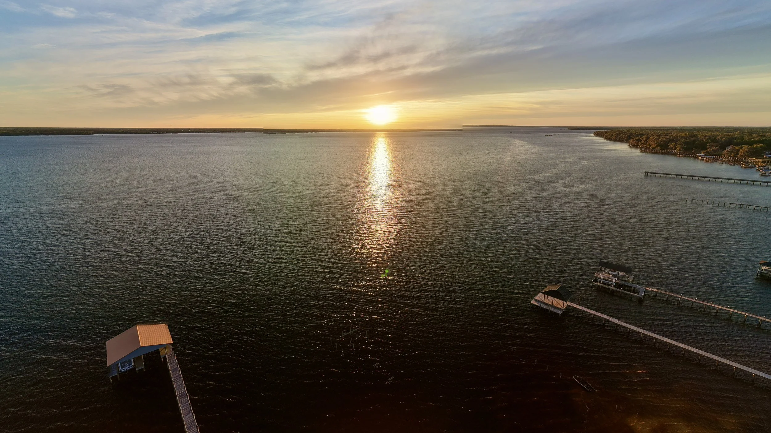 Sunset over a large body of water with several docks and small structures at the shoreline, and a partly cloudy sky.
